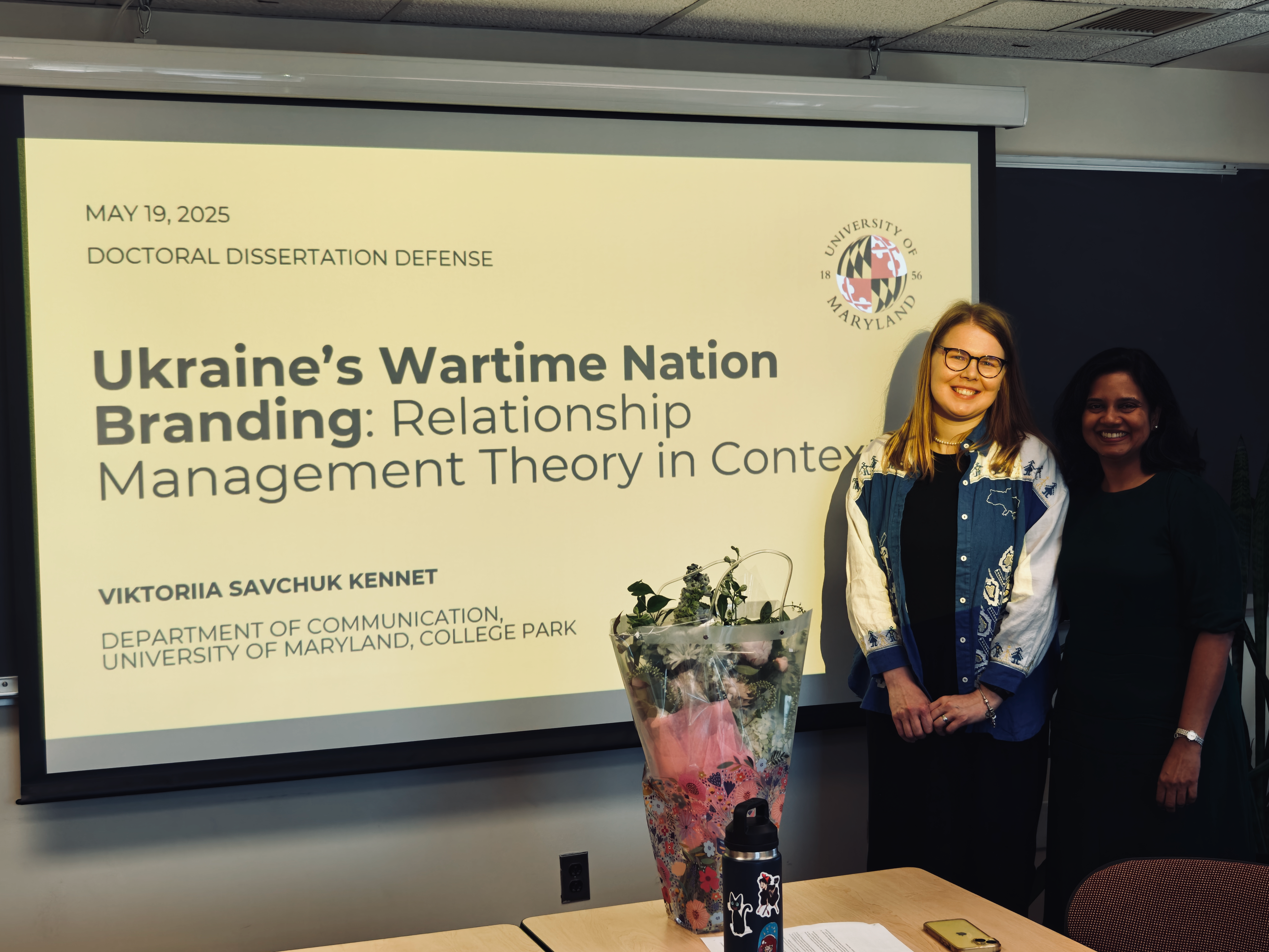 Scholar and advisor stand in front of presentation slide that reads "May 19, 2025 Doctoral Dissertation Defense, Ukraine's Wartime Nation Branding: Relationship Management Theory in Context"