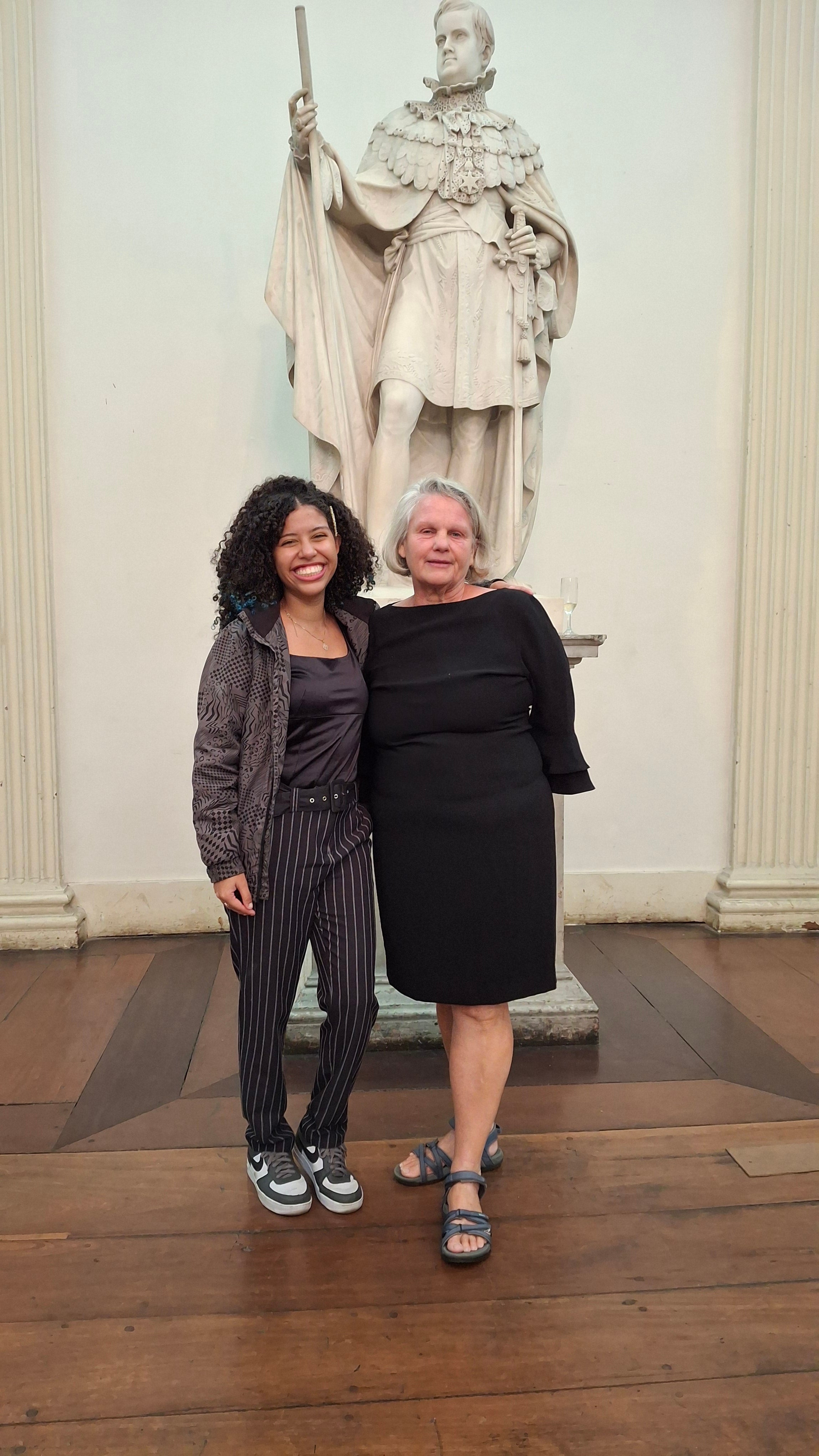 Two women stand smiling in front of a large white statue inside a historic building with wooden floors and tall columns.