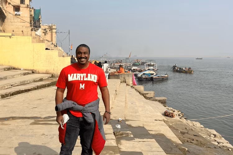 Black man in red MARYLAND shirt and black trousers stands smiling beside the Ganges river in India