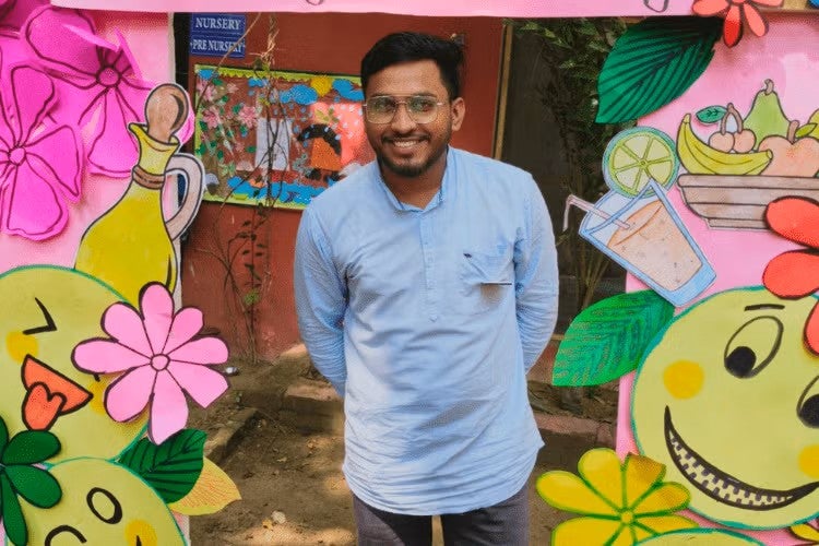 Indian man smiles with his hands behind his back, standing in between large paper flowers and smiley faces
