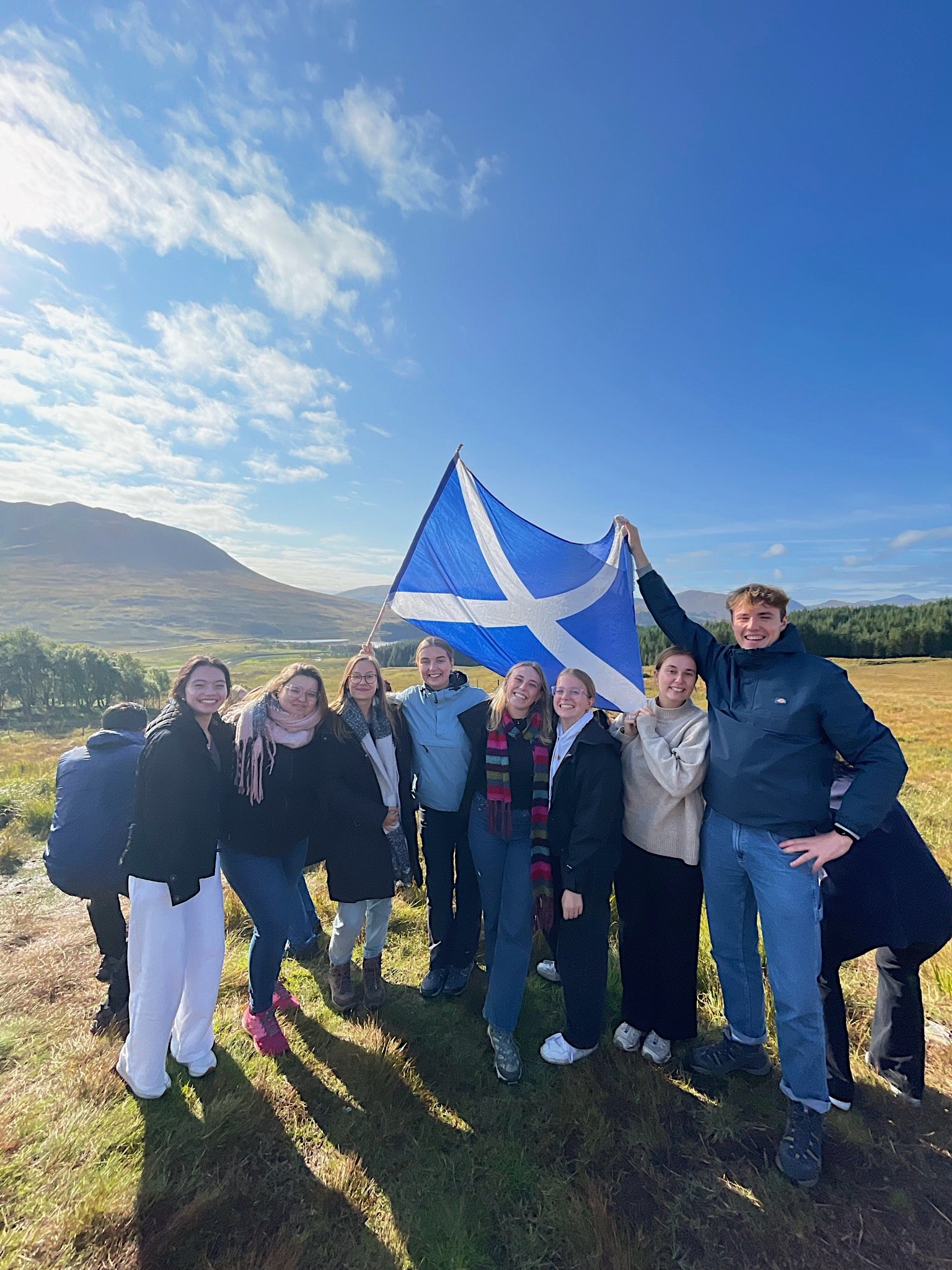 A group of students smile on a sunny day. They stand in grass and hold a Scottish flag. 