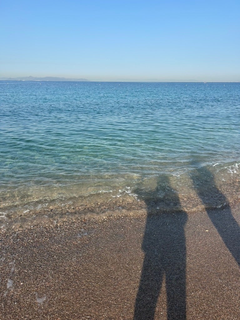 Shadows of two people standing together on a pebbled beach, looking out over clear blue water.