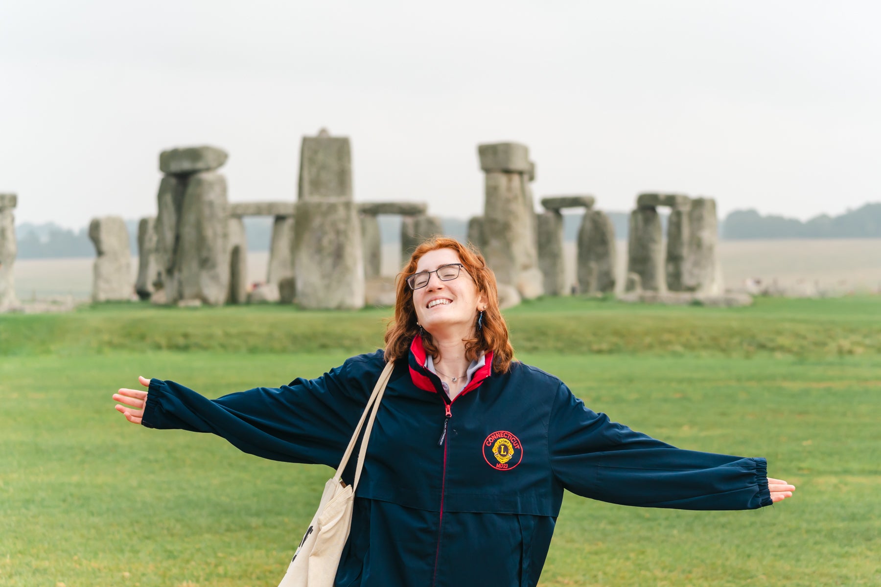 A student stands with arms outstretched in a grassy field in front of Stonehenge, smiling with the ancient stone circle in the background.