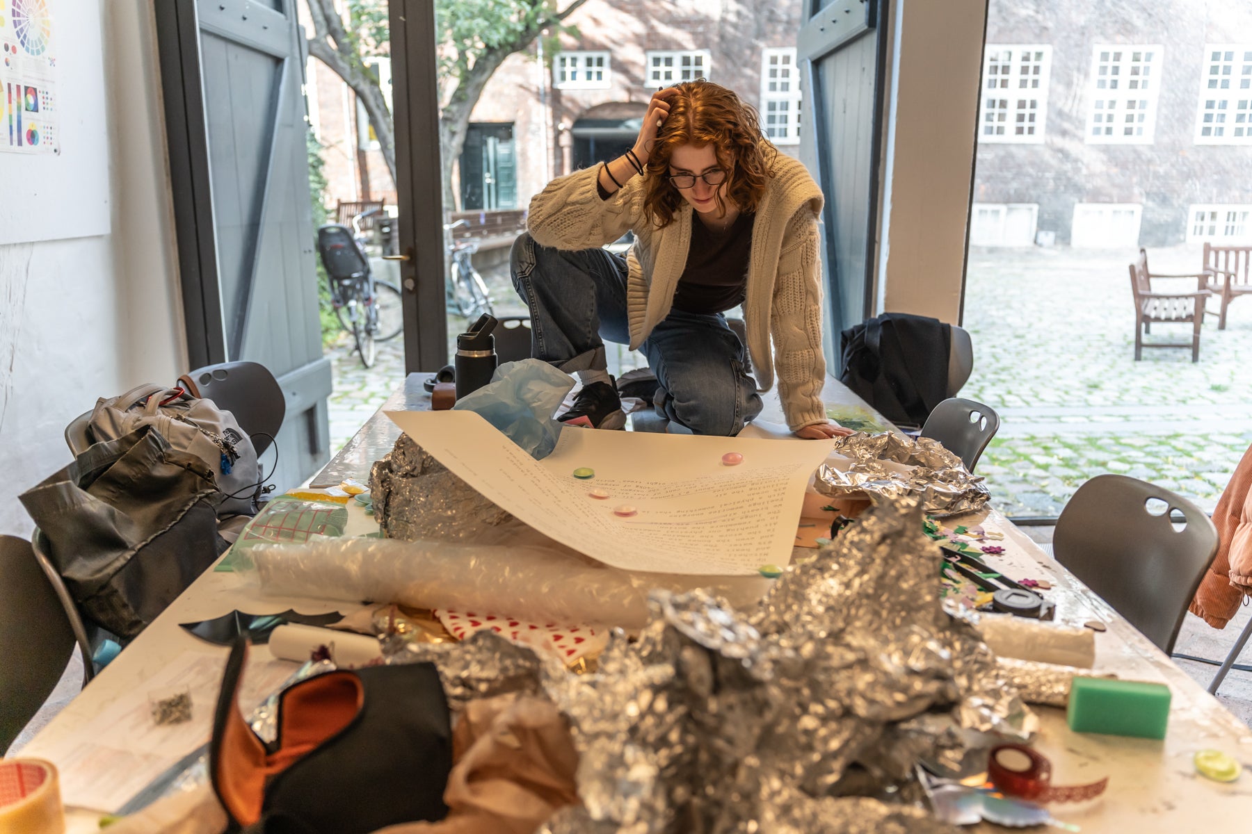 Jess leans over a cluttered table of craft materials, working on a project indoors with large open doors leading to a courtyard outside.