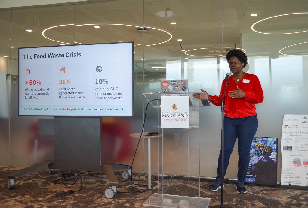A Black woman in a red top stands at a podium presenting a slide about The Food Waste Crisis