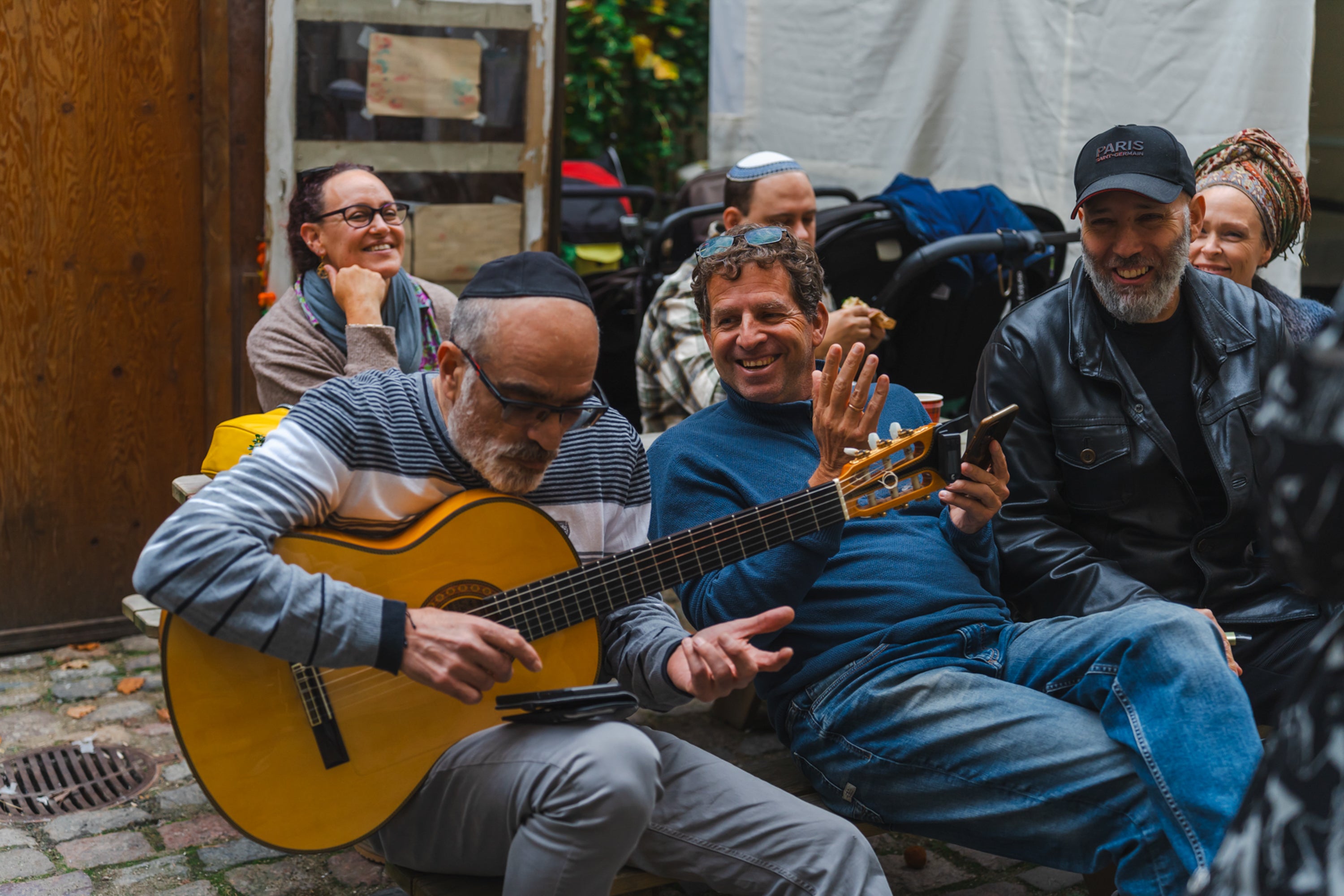 A group of adults sit outdoors laughing while one man plays a guitar, creating a relaxed, social atmosphere.