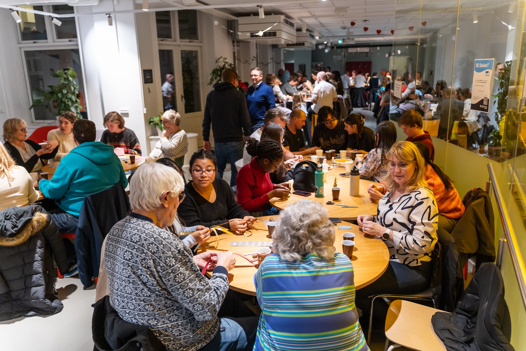 People of different ages sit around tables crafting and talking at a lively indoor community gathering.