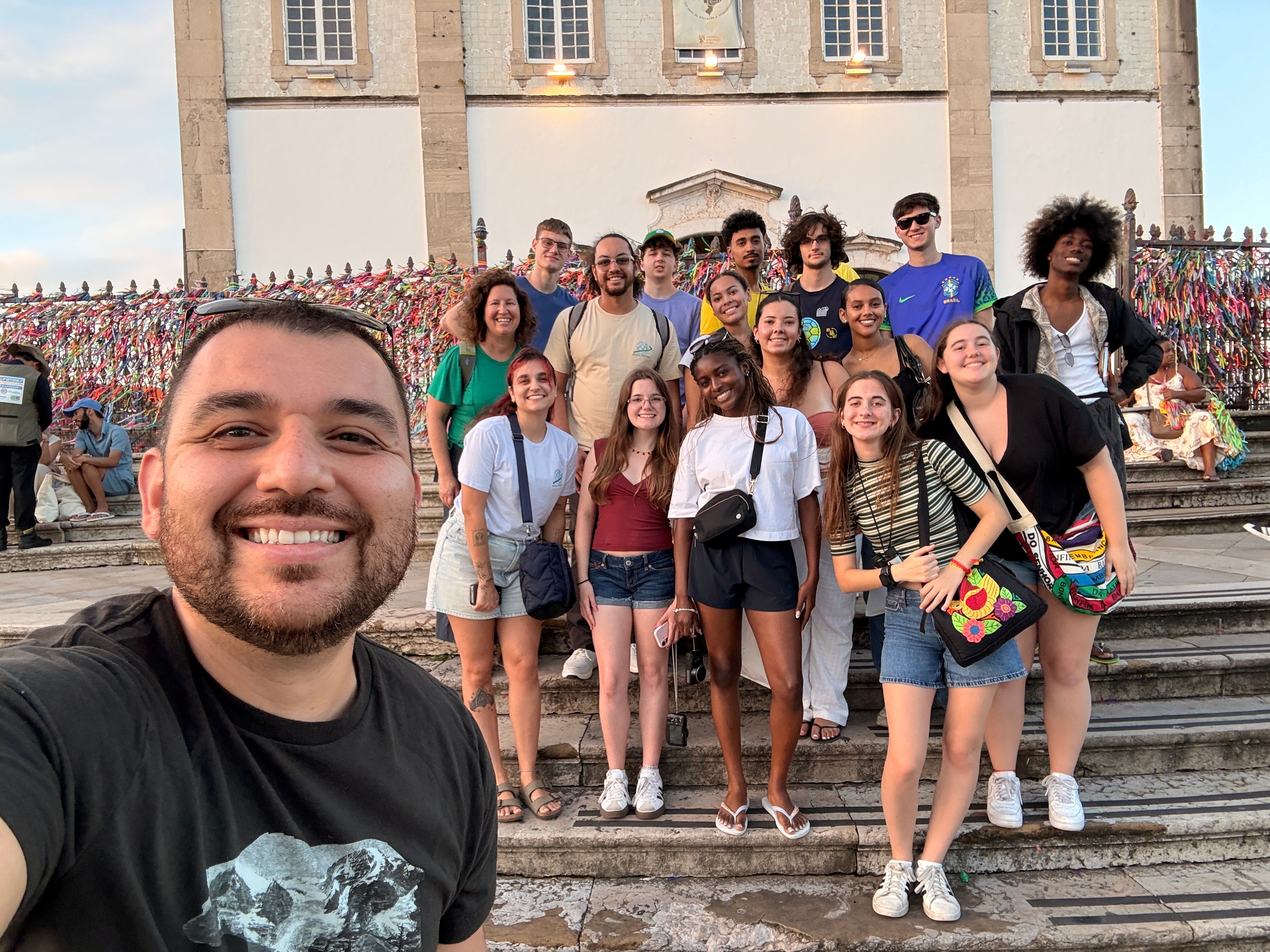 A group of students and faculty pose together on the steps of a church in front of a fence covered with colorful ribbons.