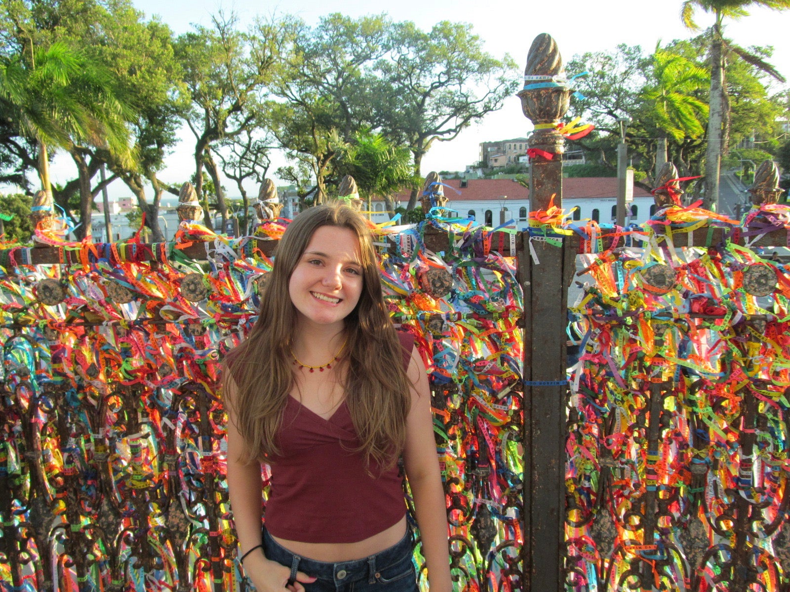 A student smiles in front of a fence densely covered with colorful ribbons, with palm trees and greenery in the background.