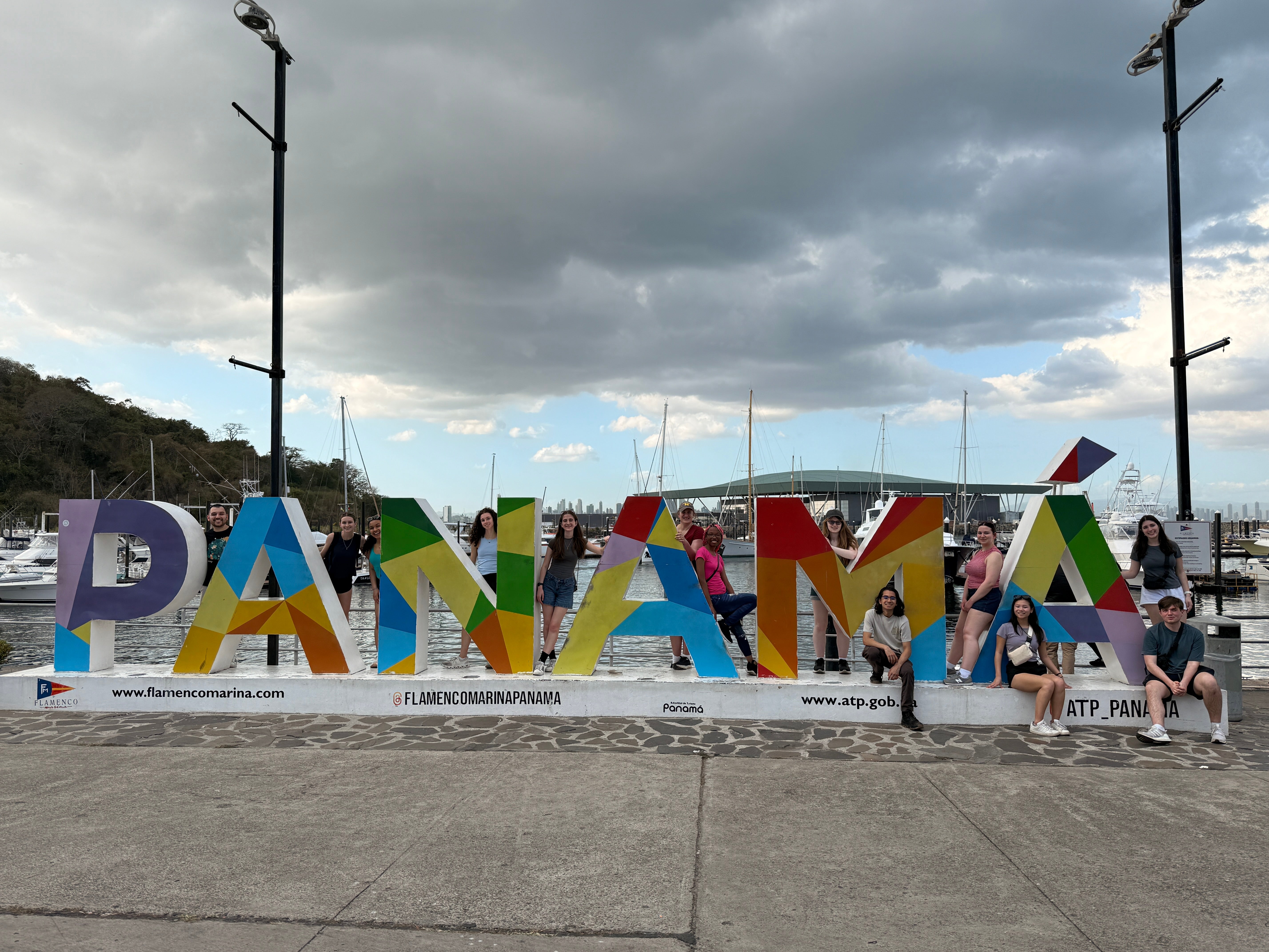 A group of students pose around large, colorful letters spelling “PANAMA” at a waterfront marina under a cloudy sky.