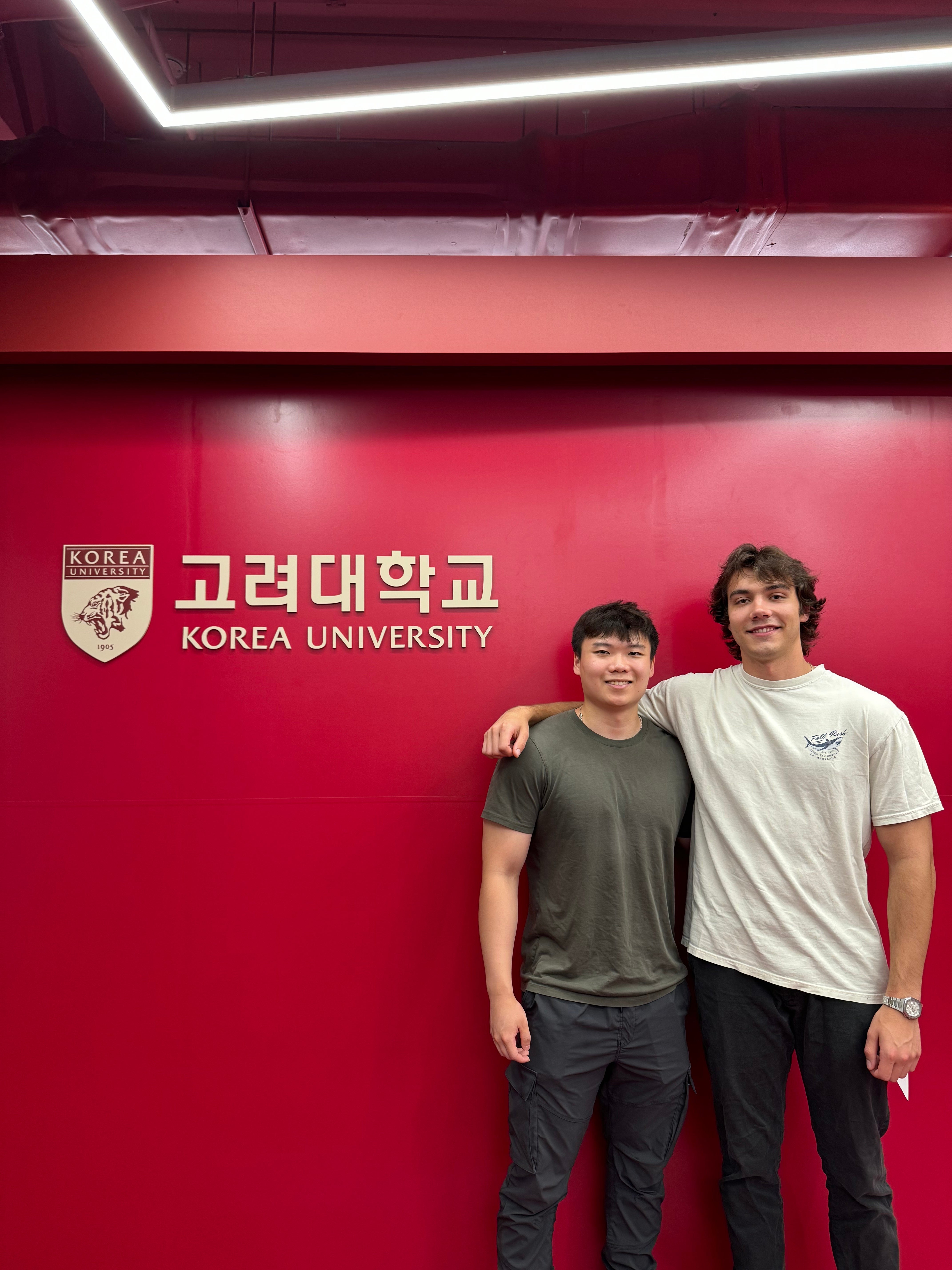 Two students stand smiling in front of a bright red wall with the Korea University logo.