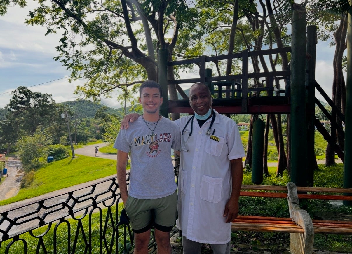 A University of Maryland student stands smiling with a local doctor wearing a white coat and stethoscope in an outdoor setting surrounded by lush greenery and trees.