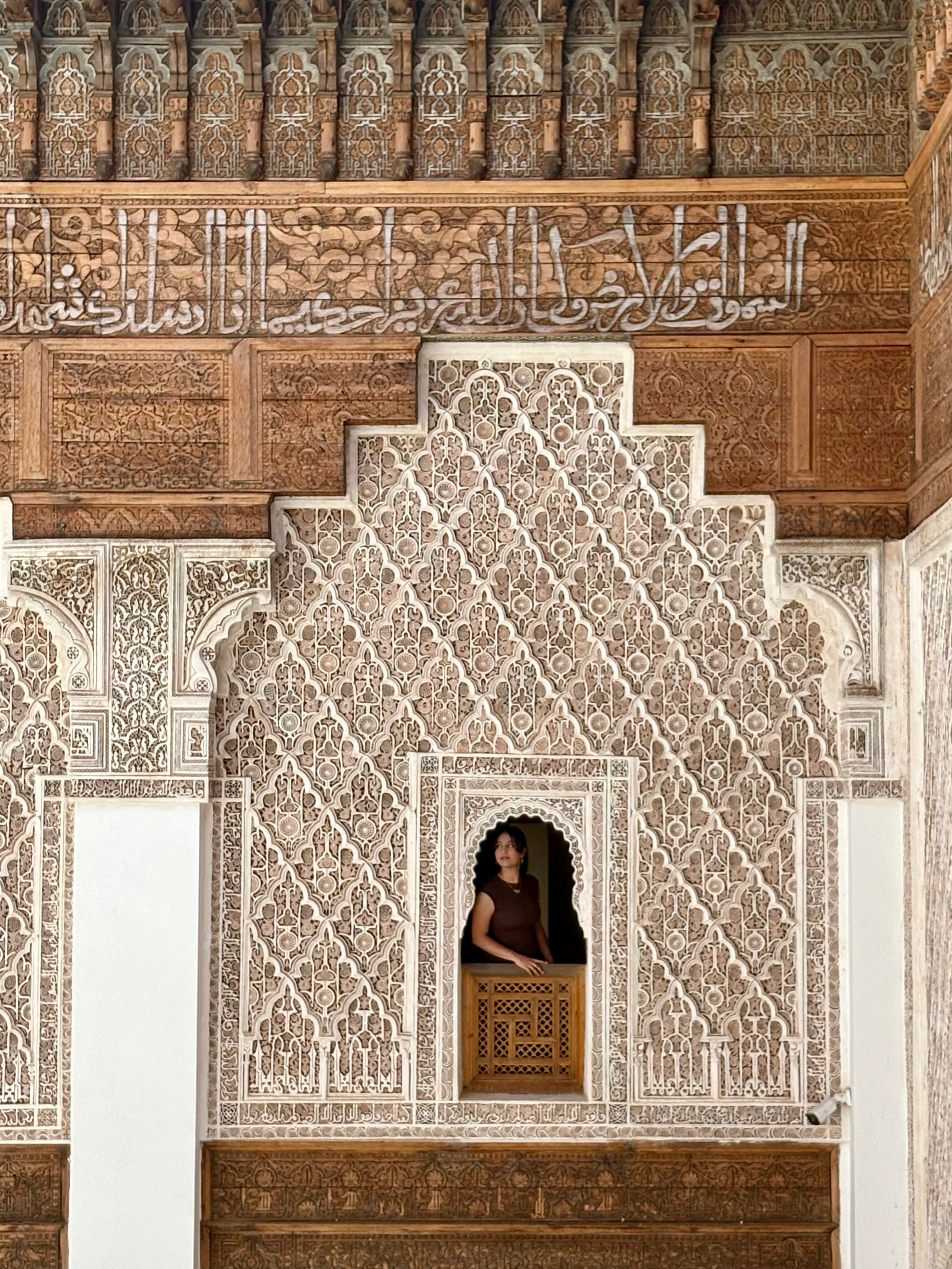 A student looks out from a carved wooden window set into an ornate wall decorated with intricate geometric patterns and Arabic calligraphy.