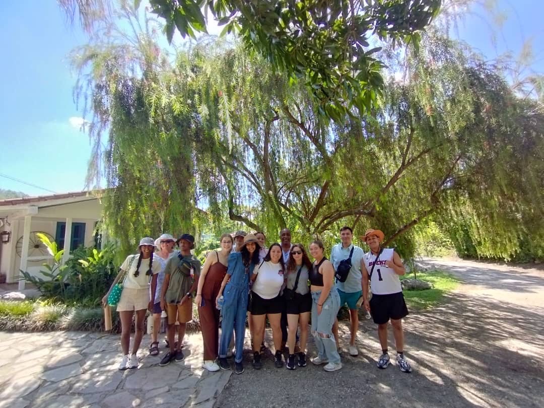 A group of students stand together smiling under a large, leafy tree in Las Terrazas, Cuba, with sunlight filtering through the branches and a small building in the background.