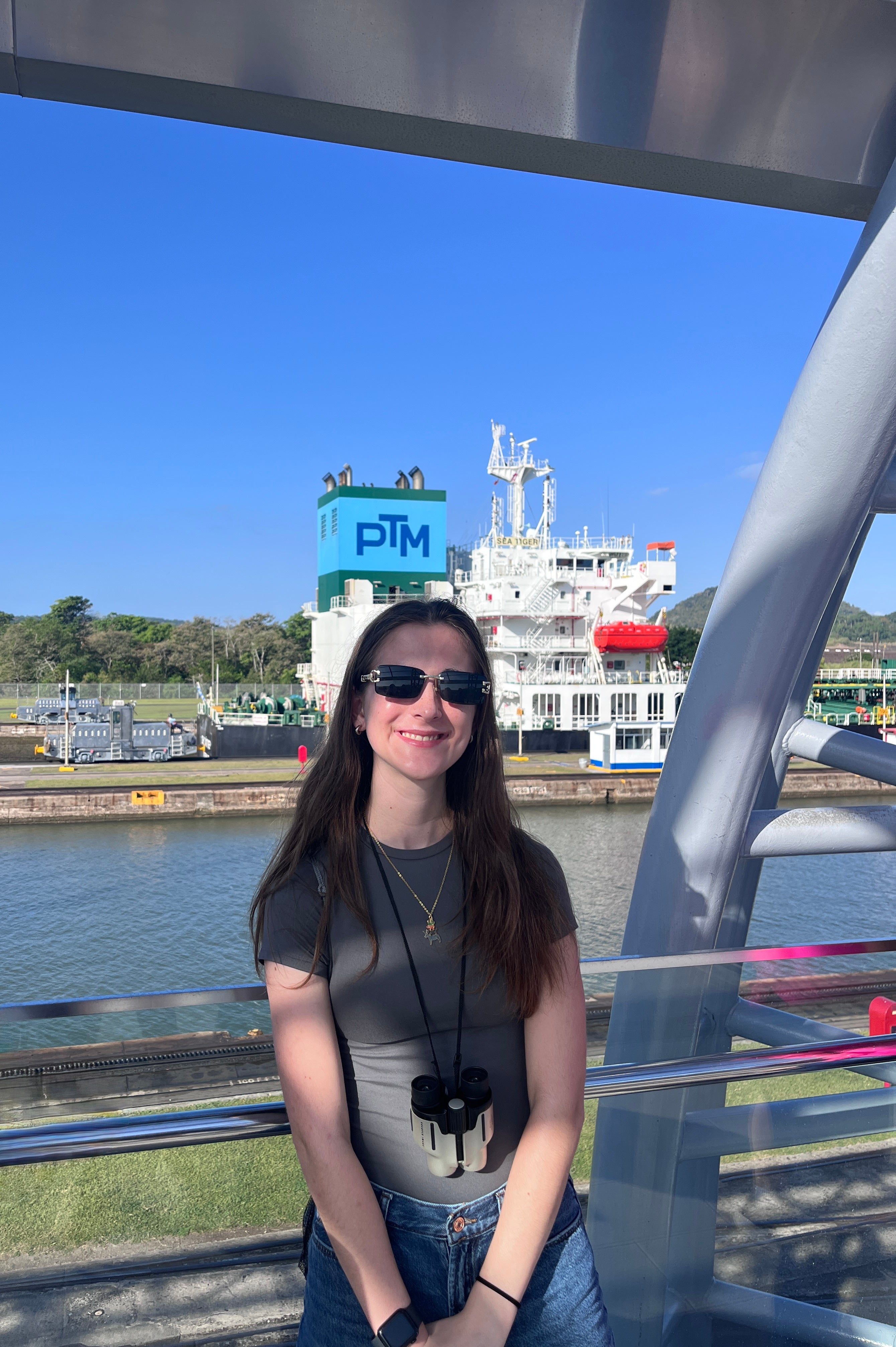 A student wearing sunglasses and binoculars smiles beside the Panama Canal, with a large ship and canal infrastructure behind her.