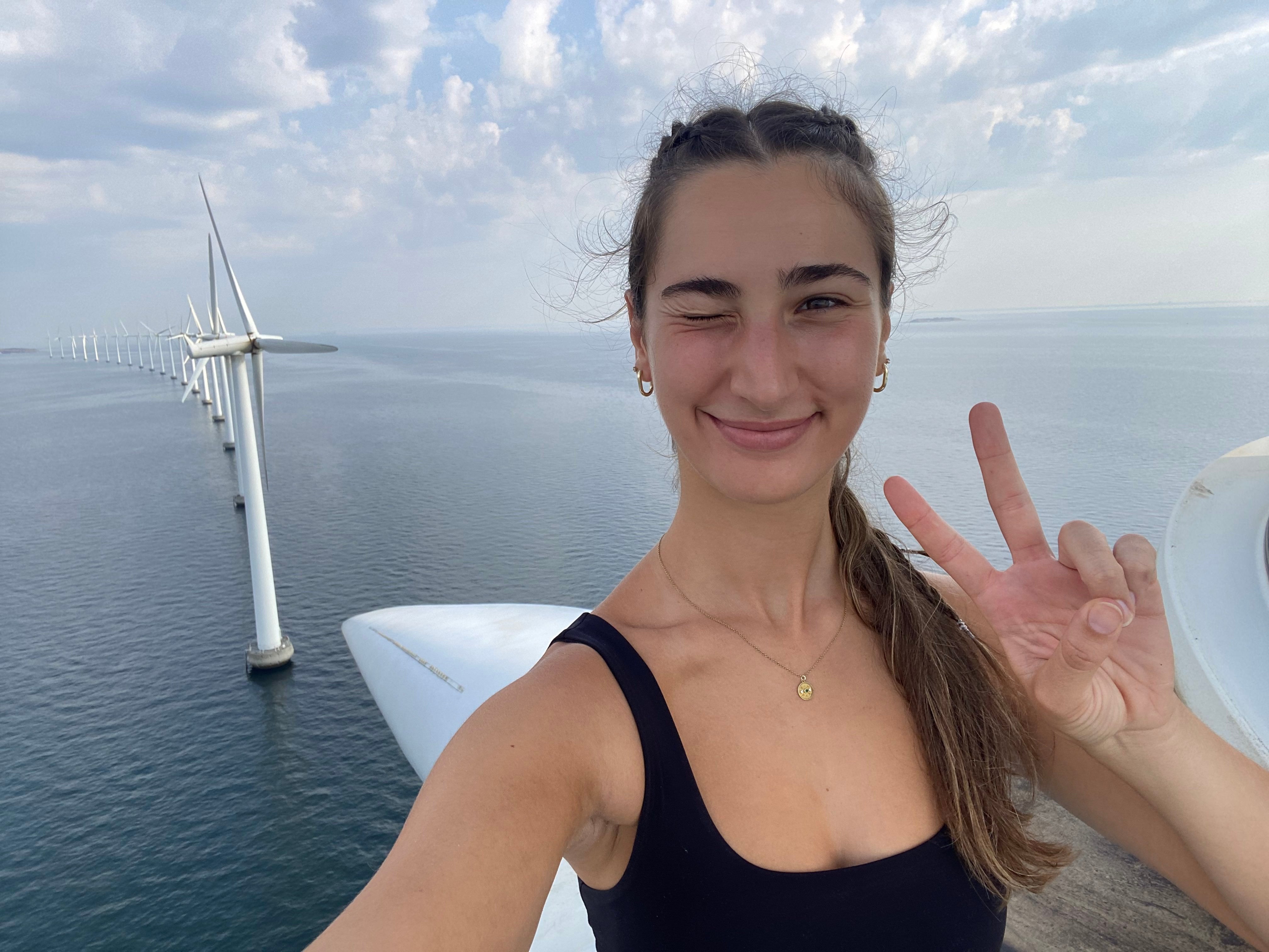 A student stands on top of a wind turbine above the sea. A wind turbine farm is behind her. 
