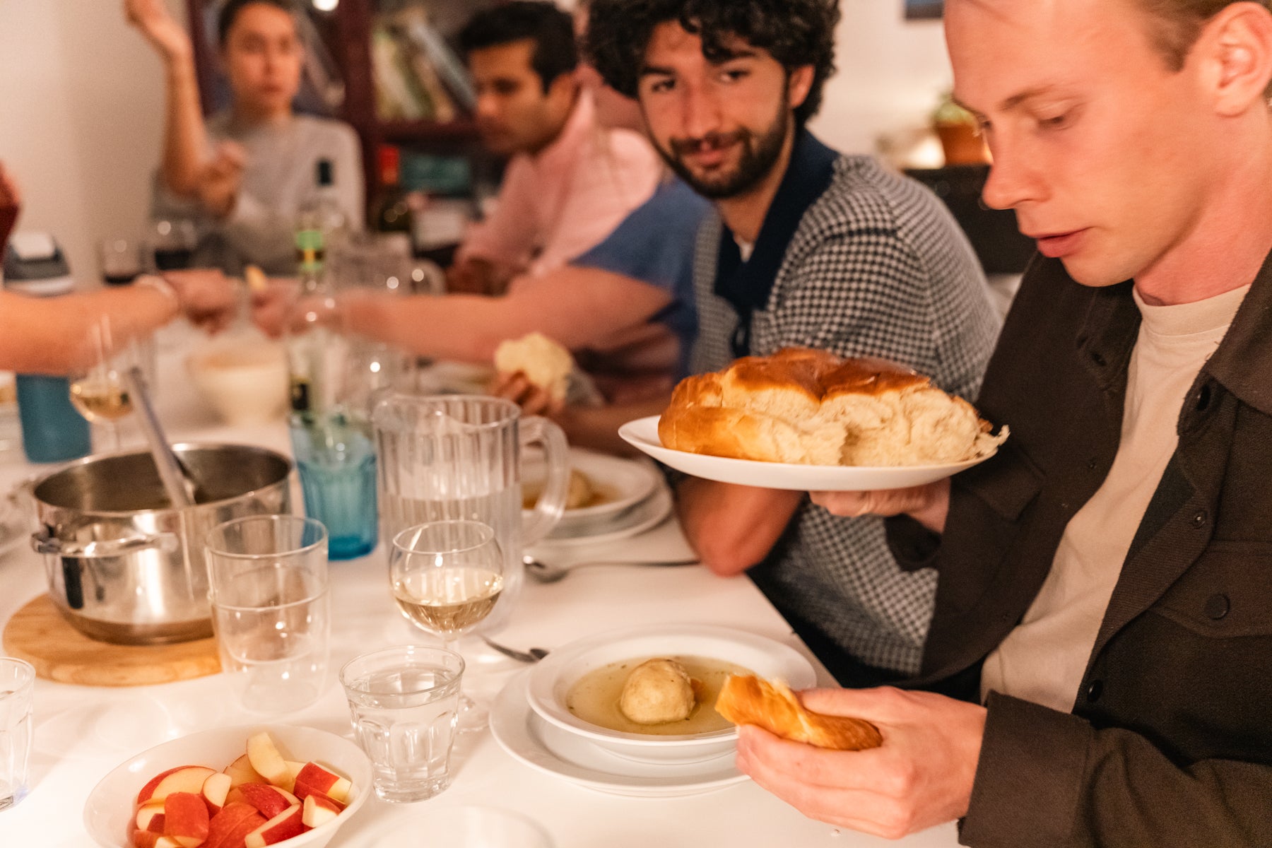 Students sit around a dining table sharing a meal, with one person holding a loaf of bread while others pass food and talk.