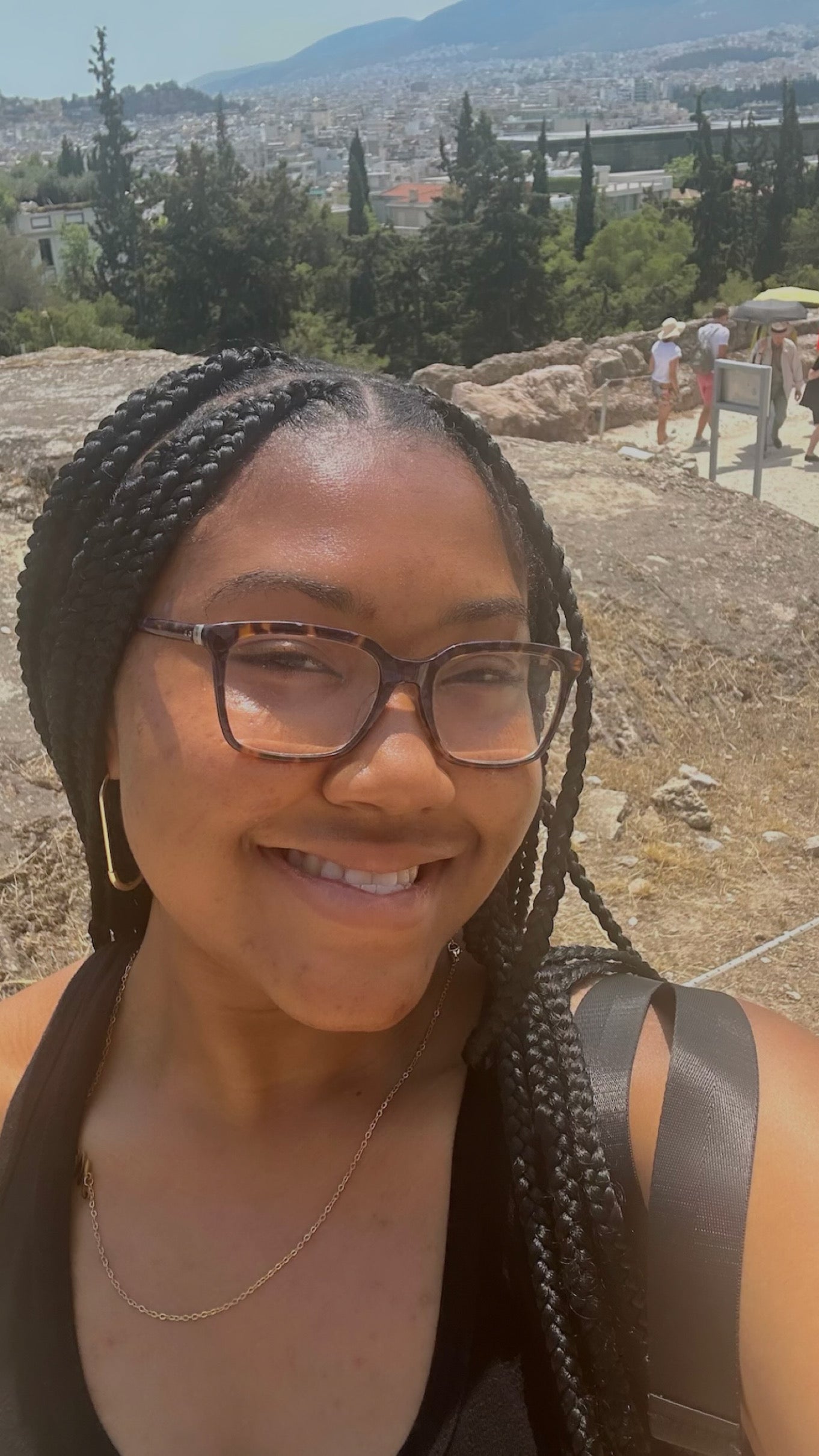 Smiling student with braided hair and glasses takes a selfie on a hillside overlooking Athens, with city buildings and greenery in the background.