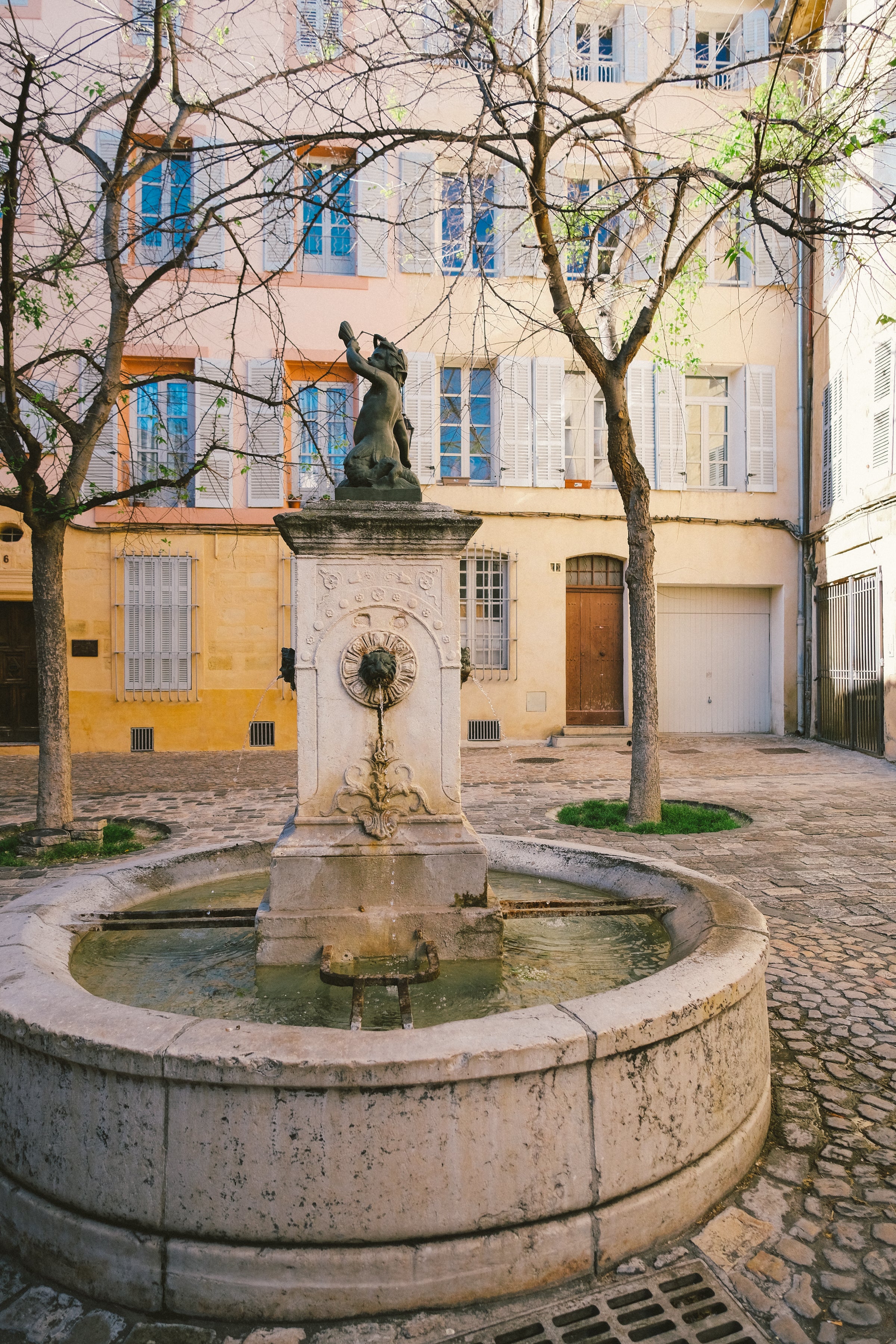 Stone fountain with a statue in a cobblestone square surrounded by pastel buildings in Aix-en-Provence.