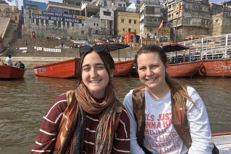 Two students smile to camera, sitting in a boat on the Ganges river with boats buildings in the background