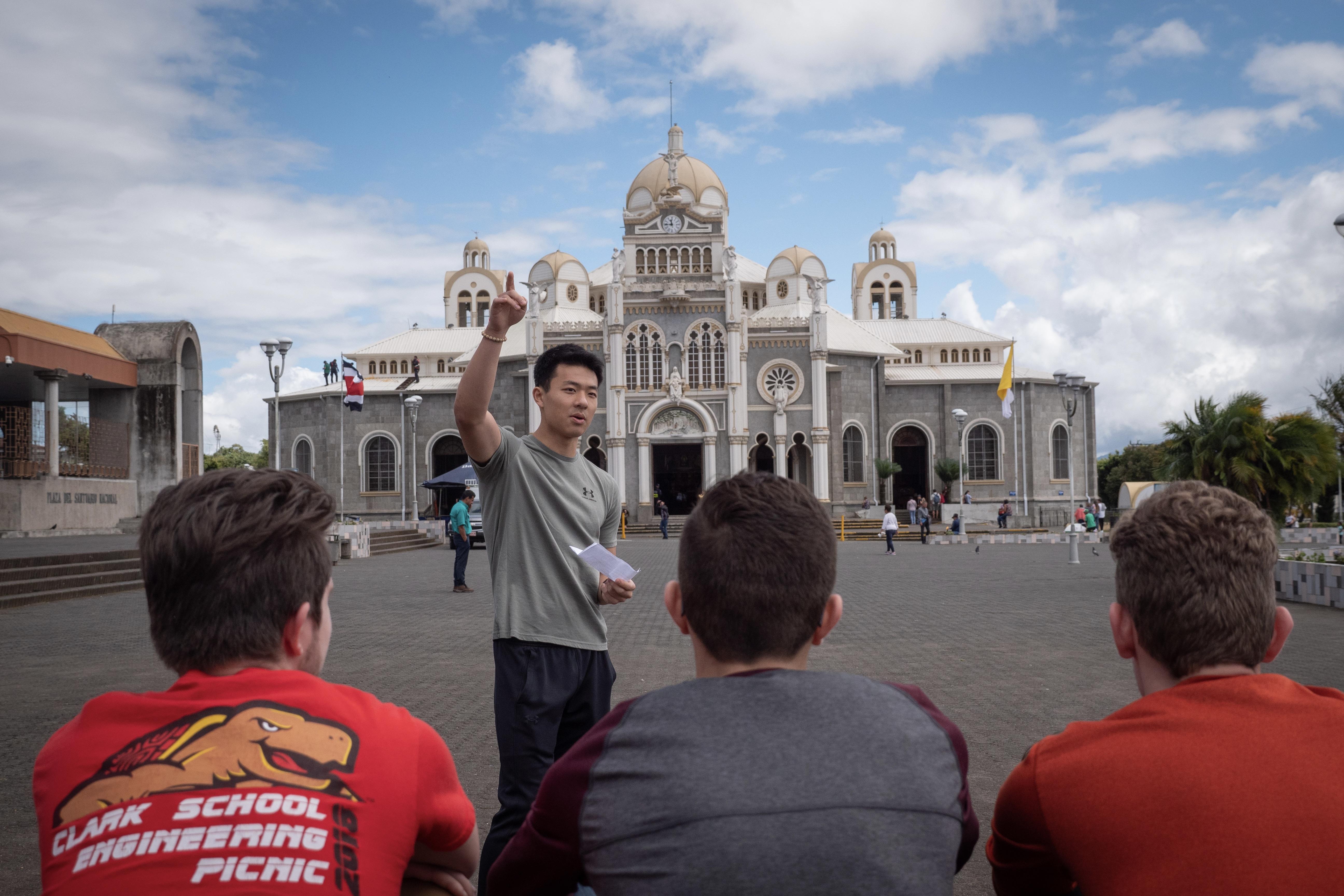 Student is giving a presentation in front of a cathedral in South America. Three other students are watching, one of whom is wearing a Clark School of Engineering t-shirt. 