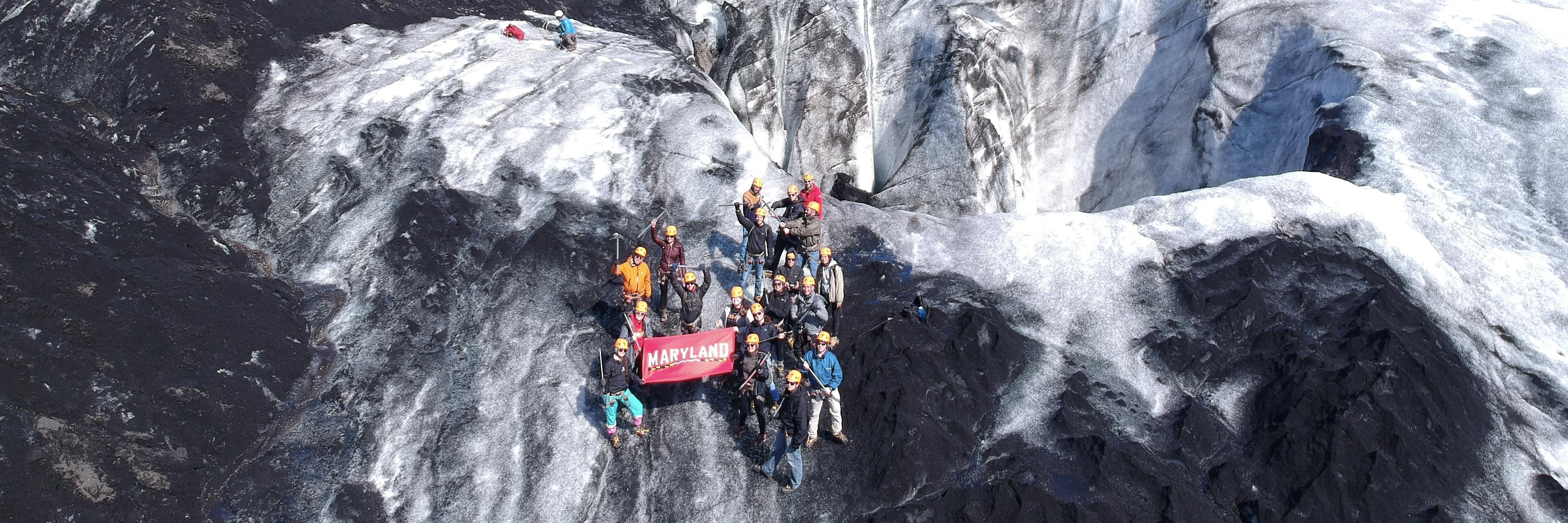 UMD students studying sustainability in Iceland take a photo with a University of Maryland flag at a glacier. 