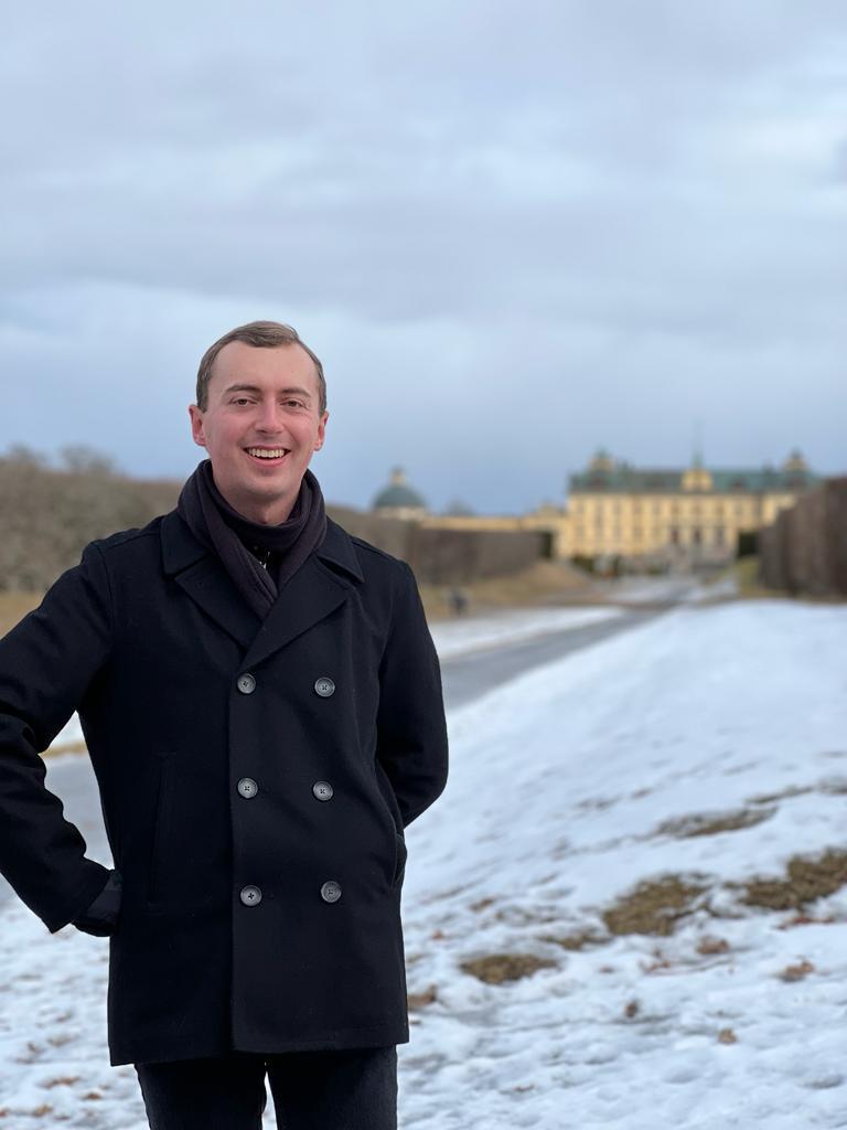 Student poses in front of a castle in Denmark. There is snow on the ground and they wear a winter coat. 