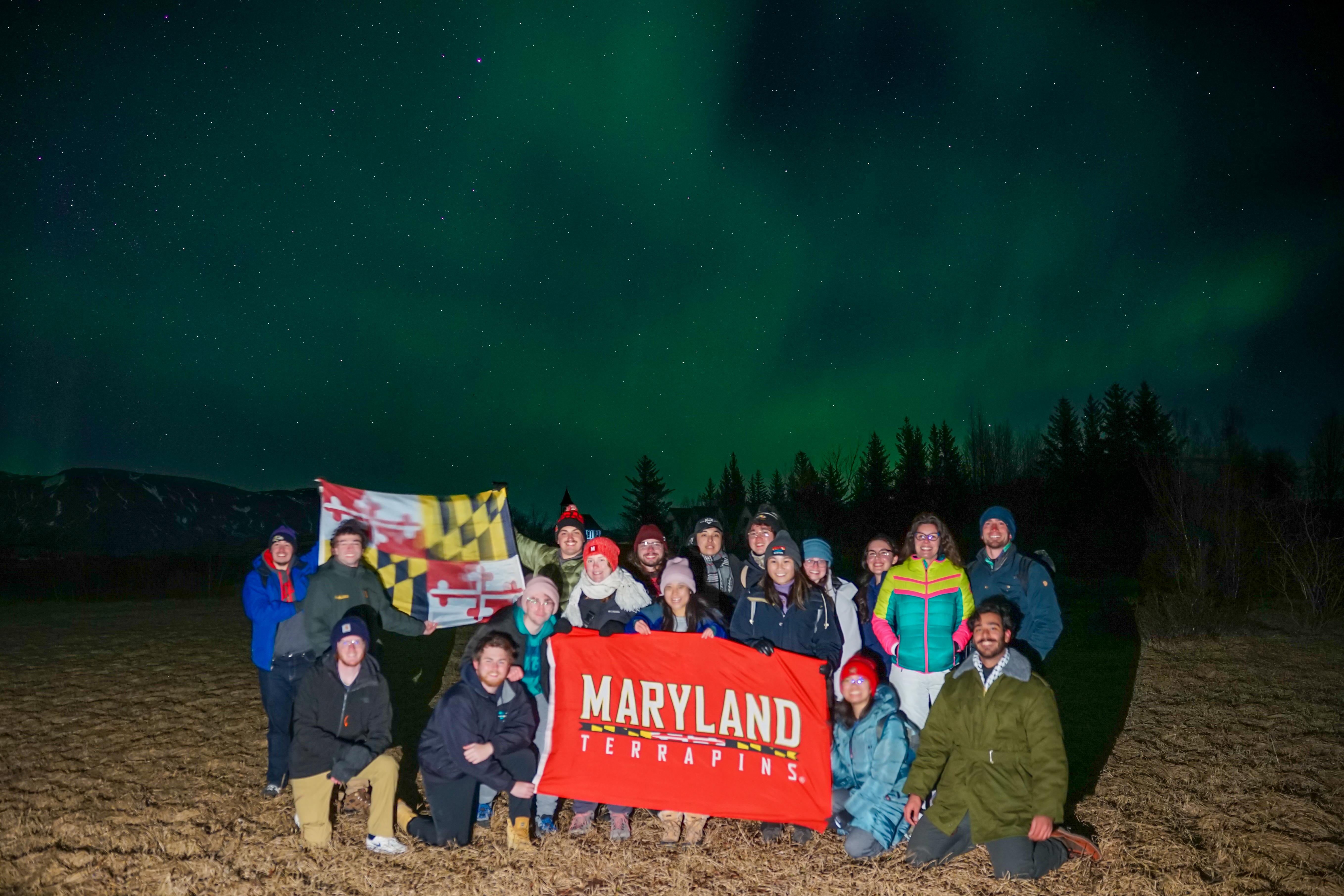 A group of students stand under the Northern lights. They hold a UMD and Maryland flag.