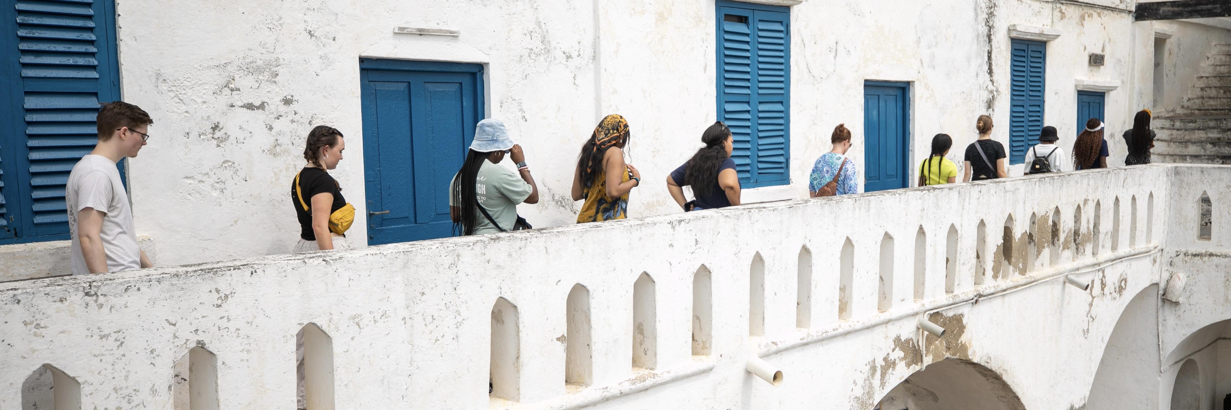 UMD students exploring Accra, Ghana. They walk along a white, stone balcony past vibrant bright doors and shutters. 