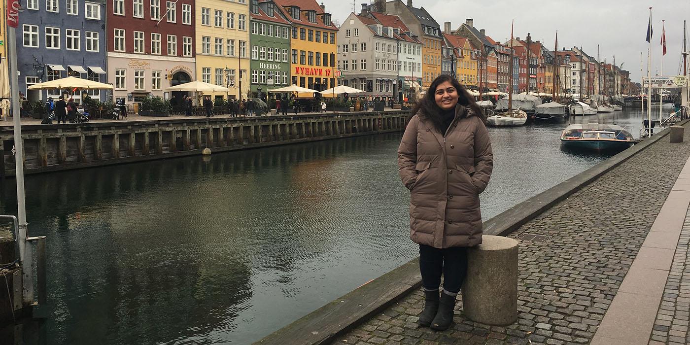 Alina in a brown winter coat, standing in front of the Nyhavn harbor in Copenhagen, Denmark.