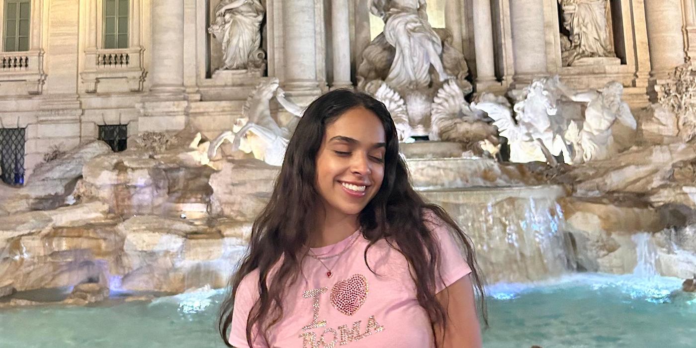 Aliya Loney in pink shirt smiling in front of the Trevi Fountain in Rome, Italy