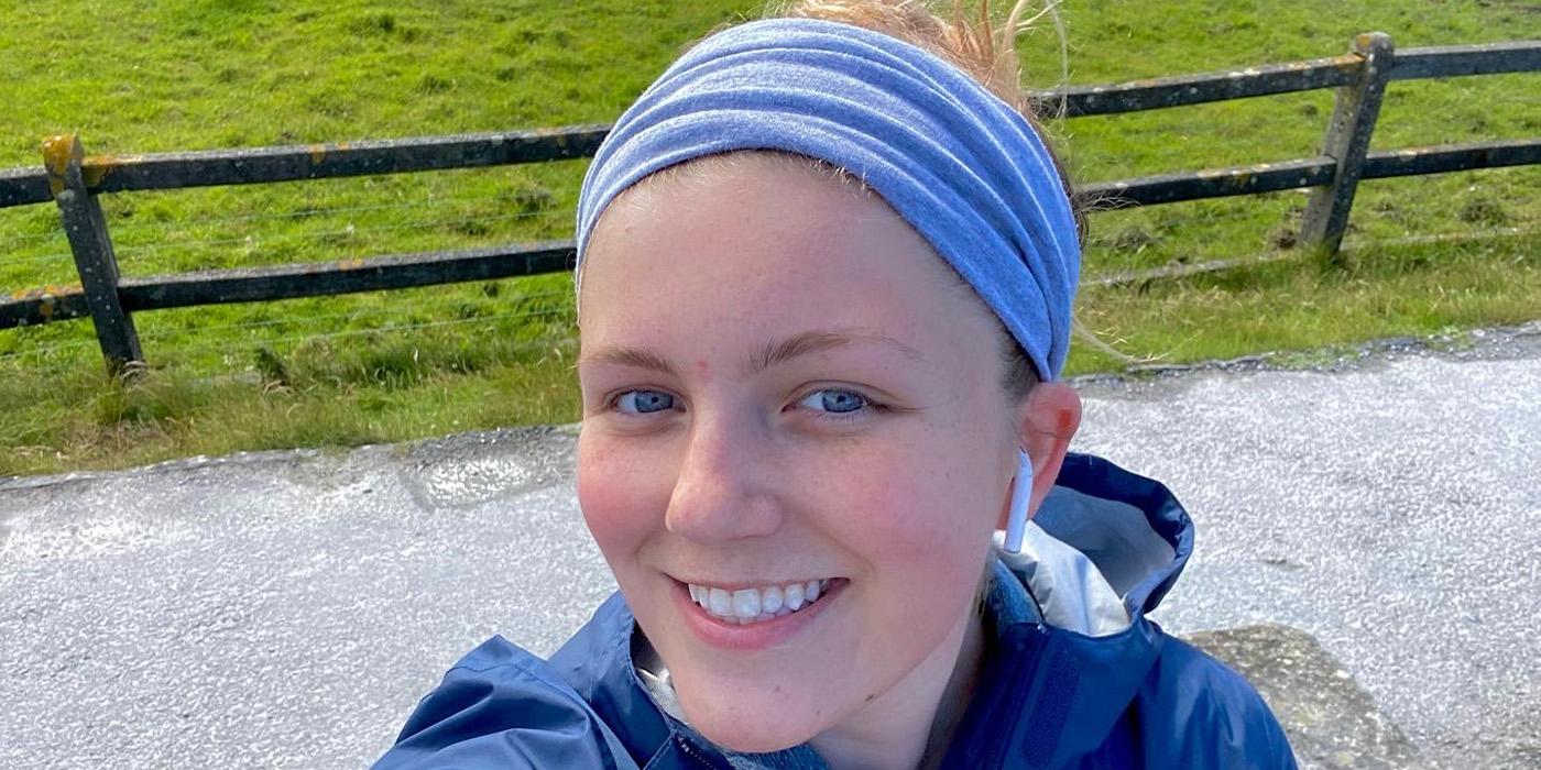 Audrey Bartholomew, wearing a light blue headband, smiles with a green field and fence behind her