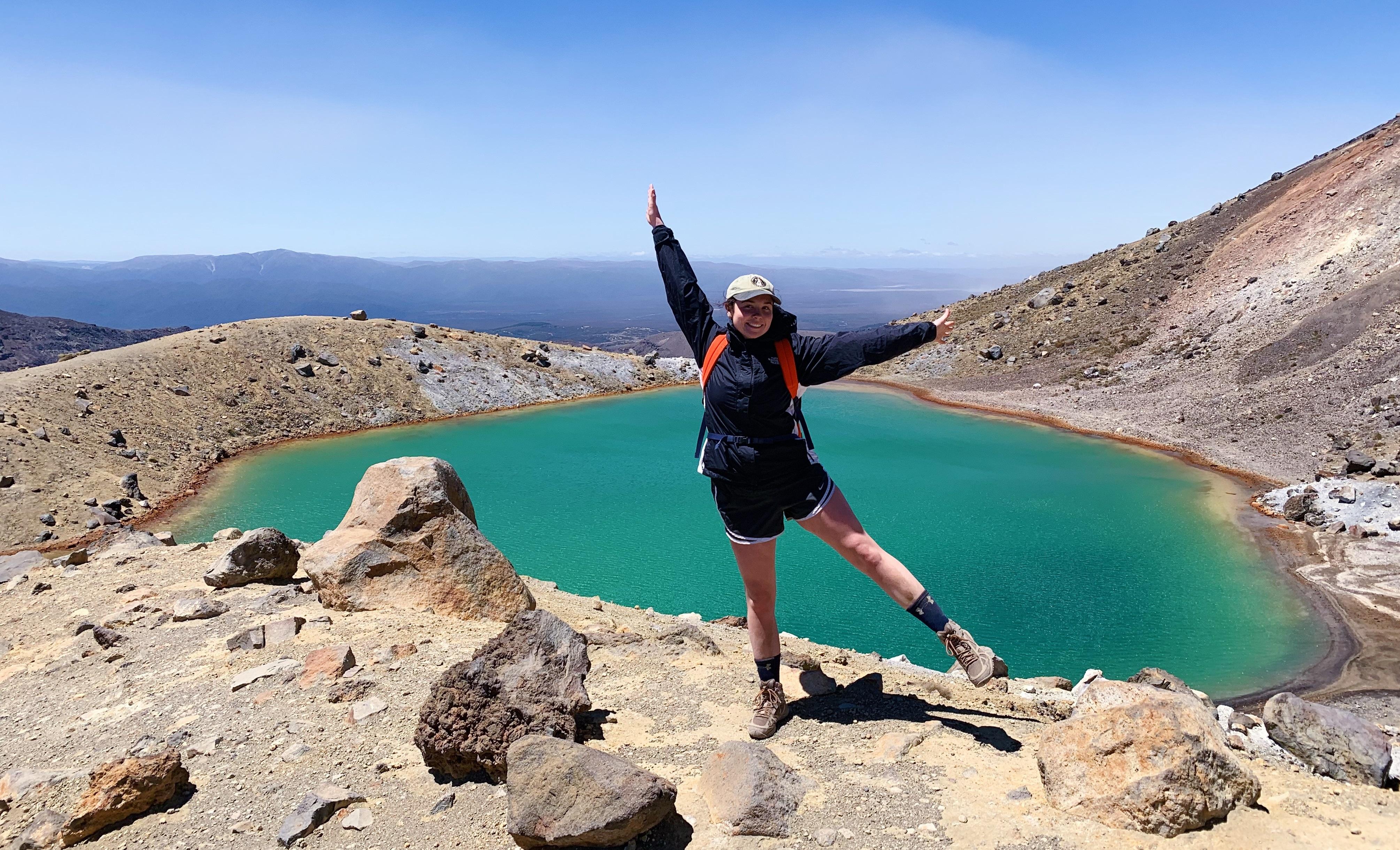 Student is standing on top of a hill with her arms raised and wide opened while a view of water is in the background