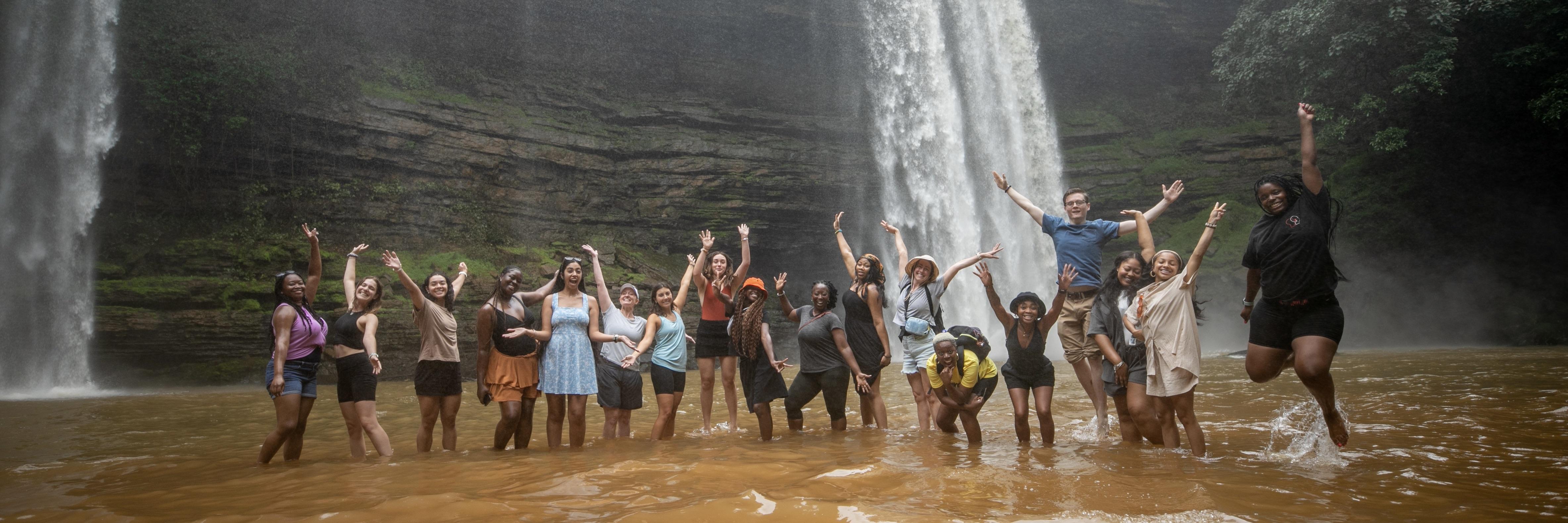 A large group of UMD students stand in a river with a flowing waterfall in the background.
