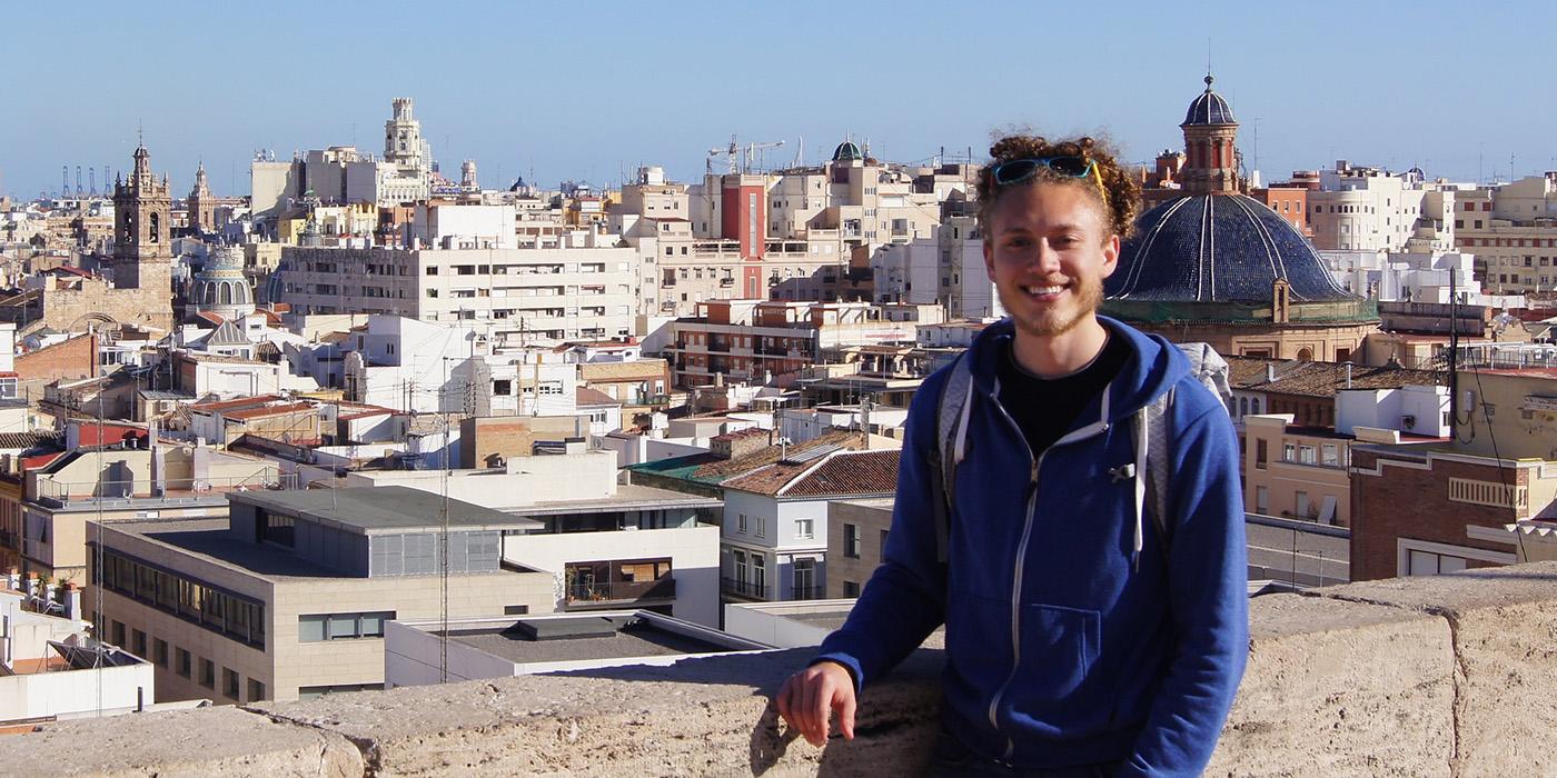 Joshua standing in front of the Valencia city skyline. He is wearing a blue jacket and backpack. 