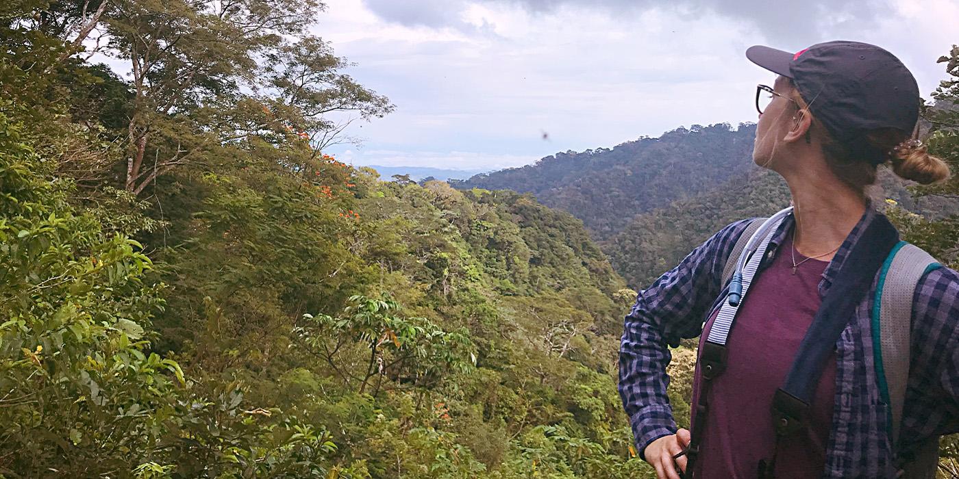 Kaitlyn Lee wears a baseball cap and looks off into the distance of a heavily forested area