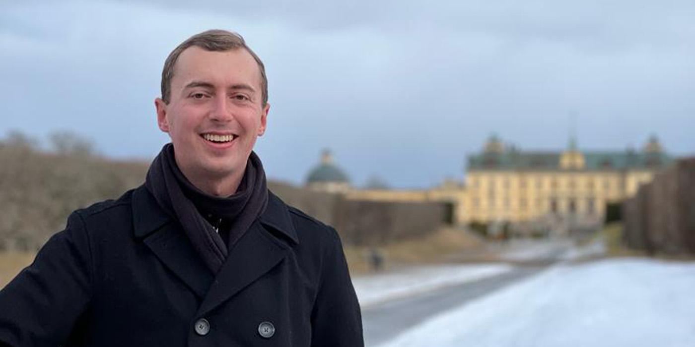 Logan Glauser, in a black pea coat, smiling in front of the Kronborg Castle in Demark 