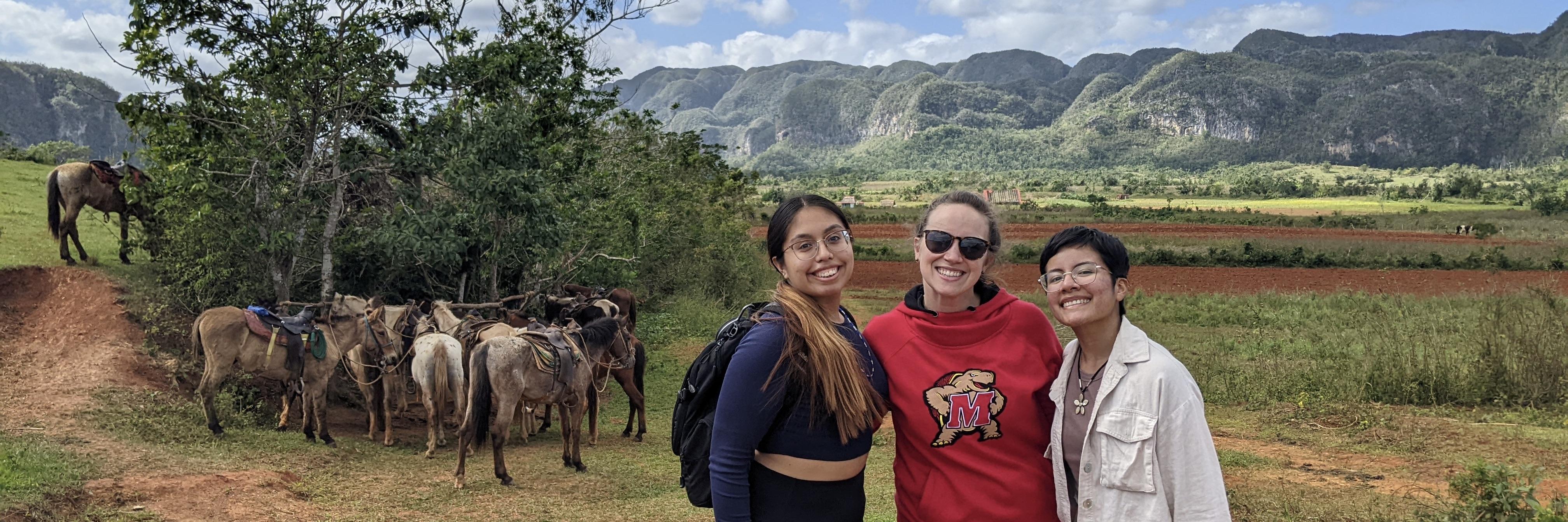 Three students stand in front of a group of horses. Green rolling hills are seen in the background and the student in the center wears a red Maryland sweatshirt with Testudo on the chest.