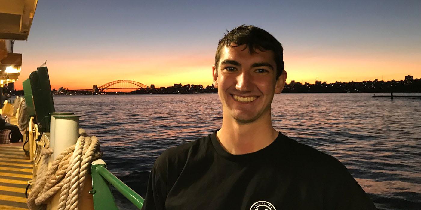 Nick smiling on a ferry boat in Sydney, Australia, with water and a bridge behind him. 