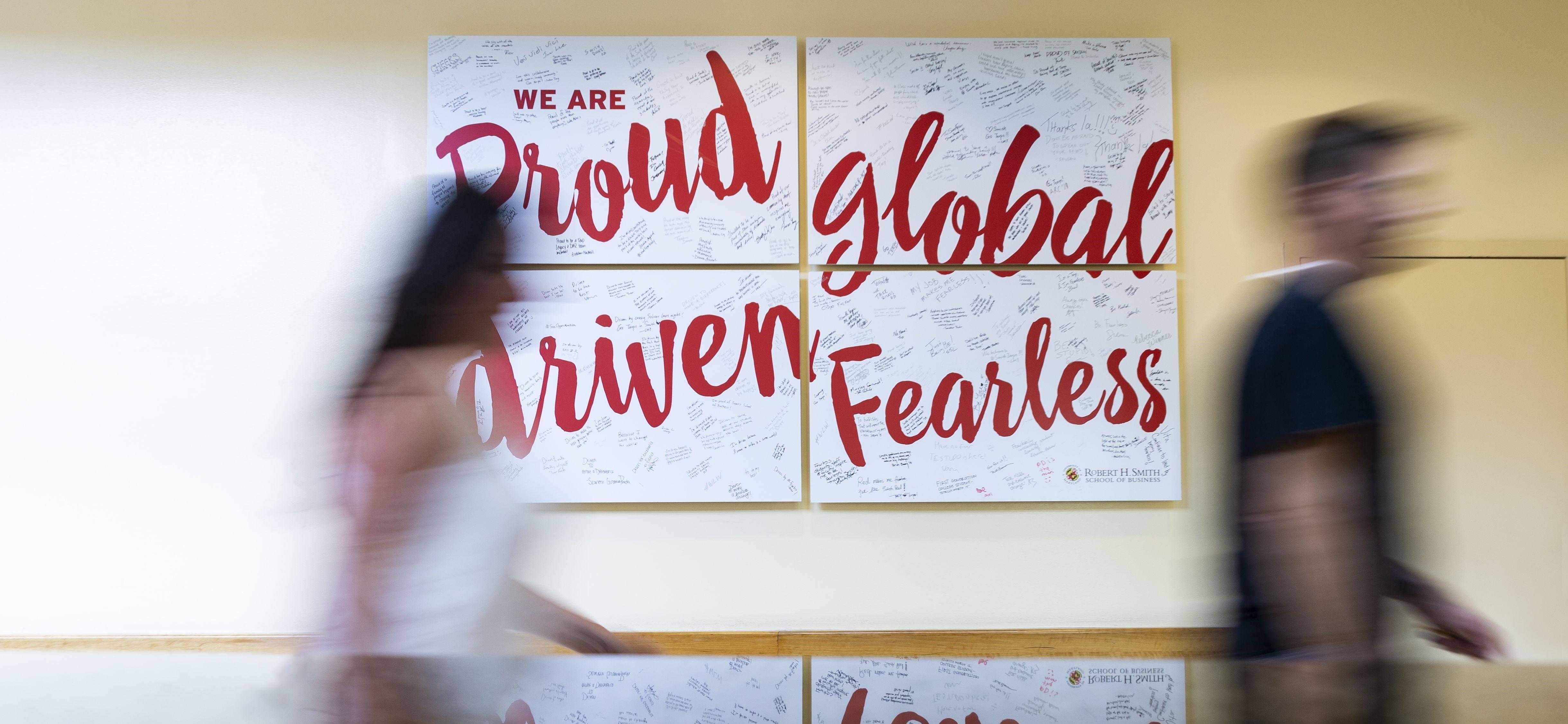 Blur of students walking in front of sign: "We are Proud, Global, Driven, Fearless" -- interior view of Van Munching Hall