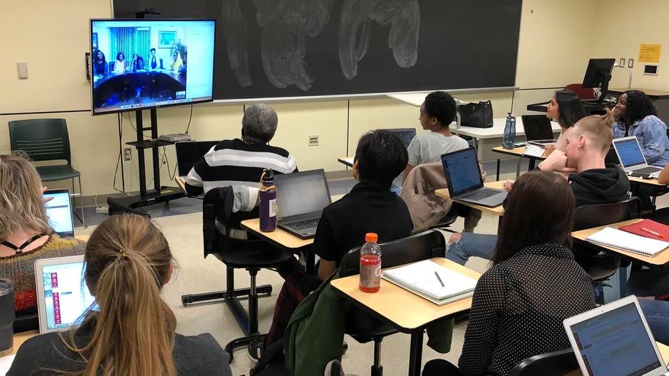 Students sit at desks facing a video conference screen at the front of the classroom during a Global Classrooms course.