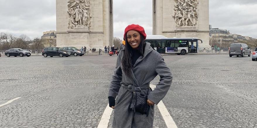 Kaya Benitez stands in front of a monument in Spain. She wears a grey coat and red beret 