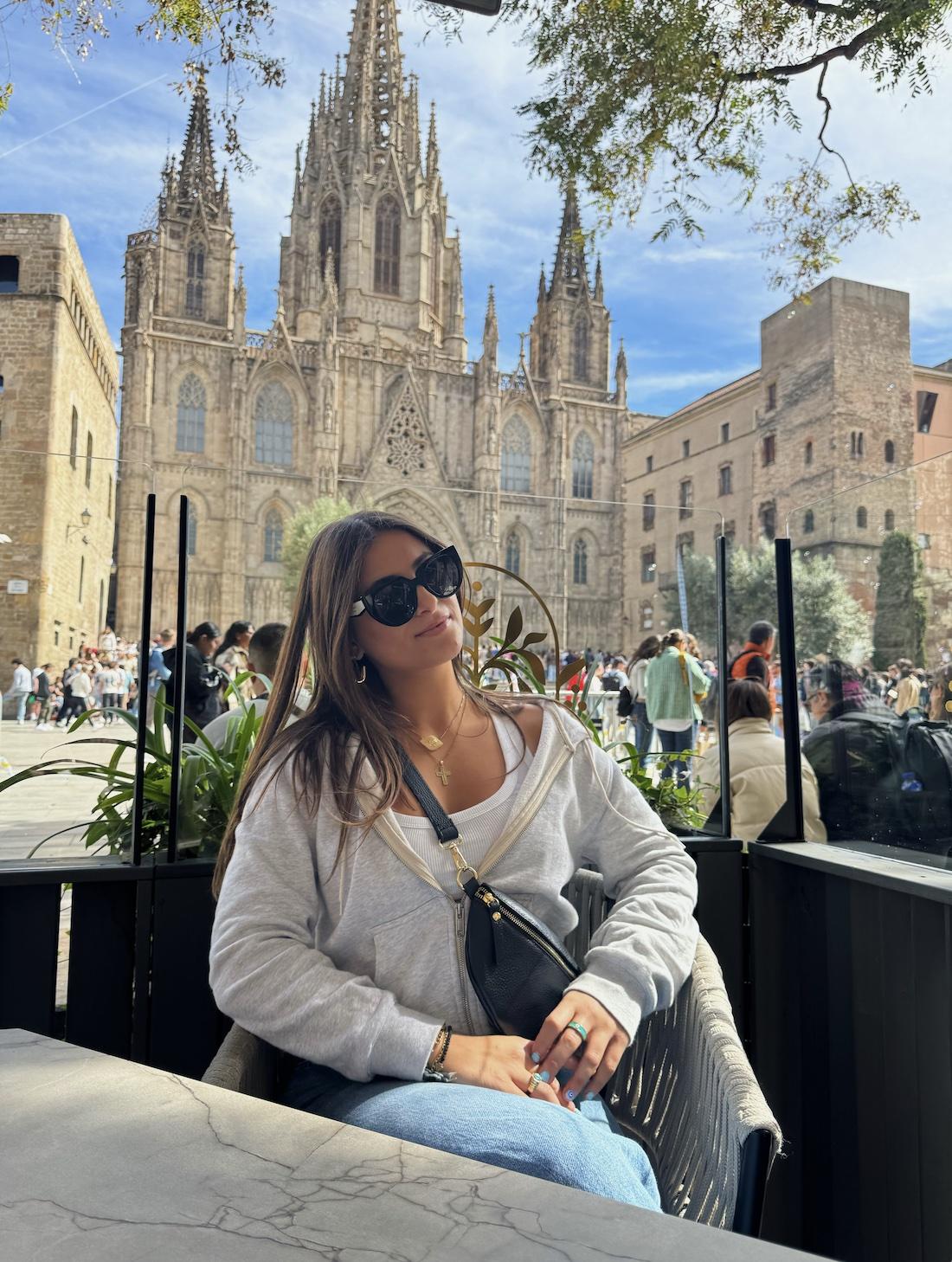 Kalliope sits at a table in Spain. A intricate Spanish church is in the background.