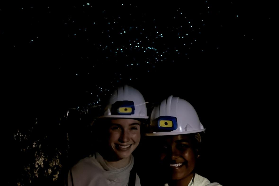 Ru and friend visit the glowworm caves in New Zealand. They stand in the dark with hard hats and bioluminescent dots are above their head.