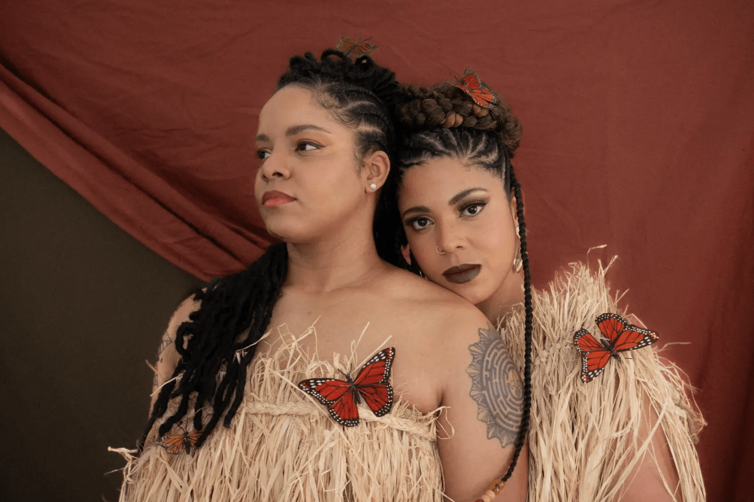 Two black women with long braids stand together. They wear garments of straw and are adorned with three butterflies.