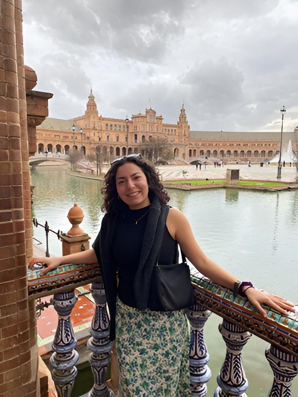 Anna stands on a stone balcony overlooking the water. Red clay buildings are on the other side of the water. 