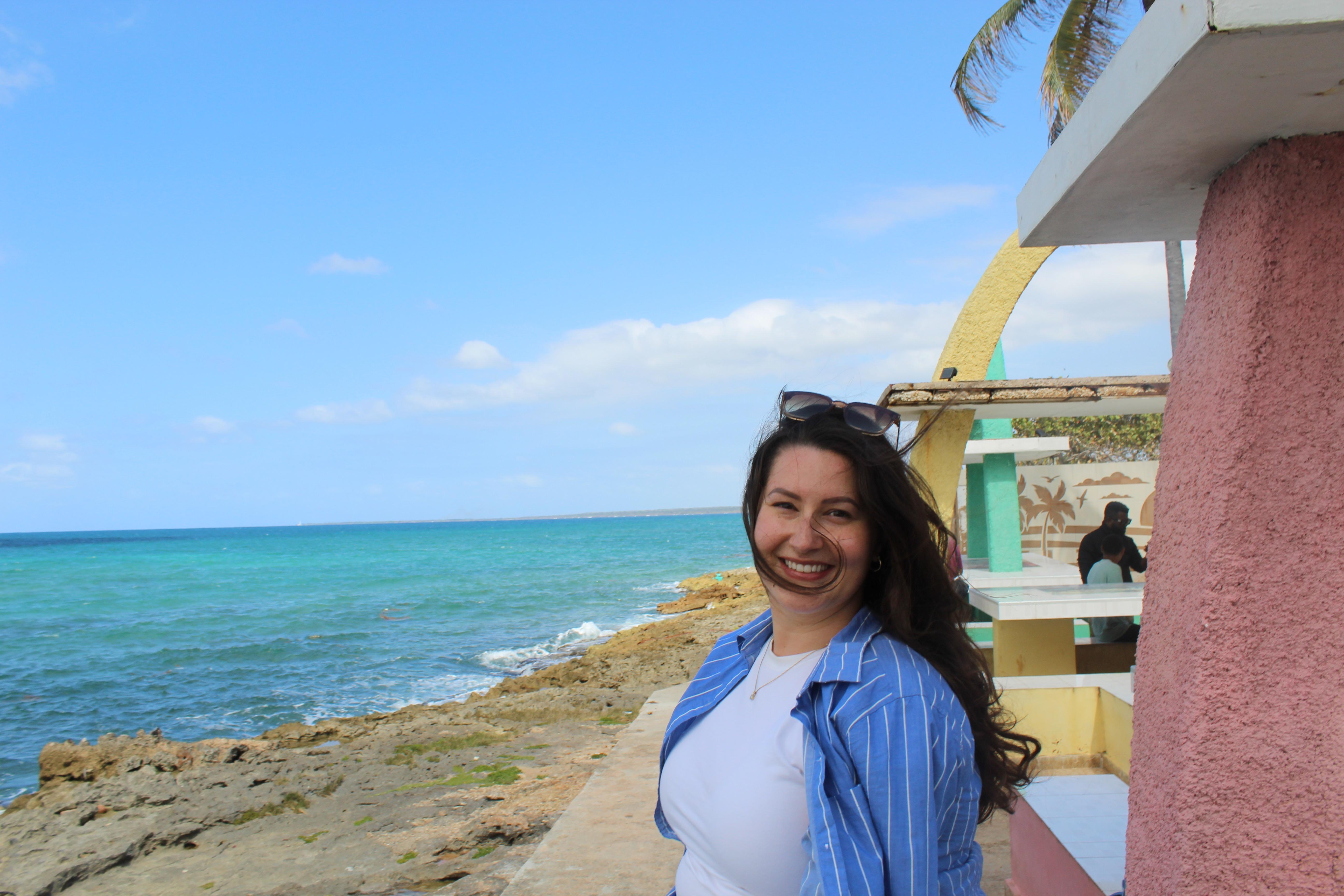 A woman with brown hair stands above a beach with clear blue water.