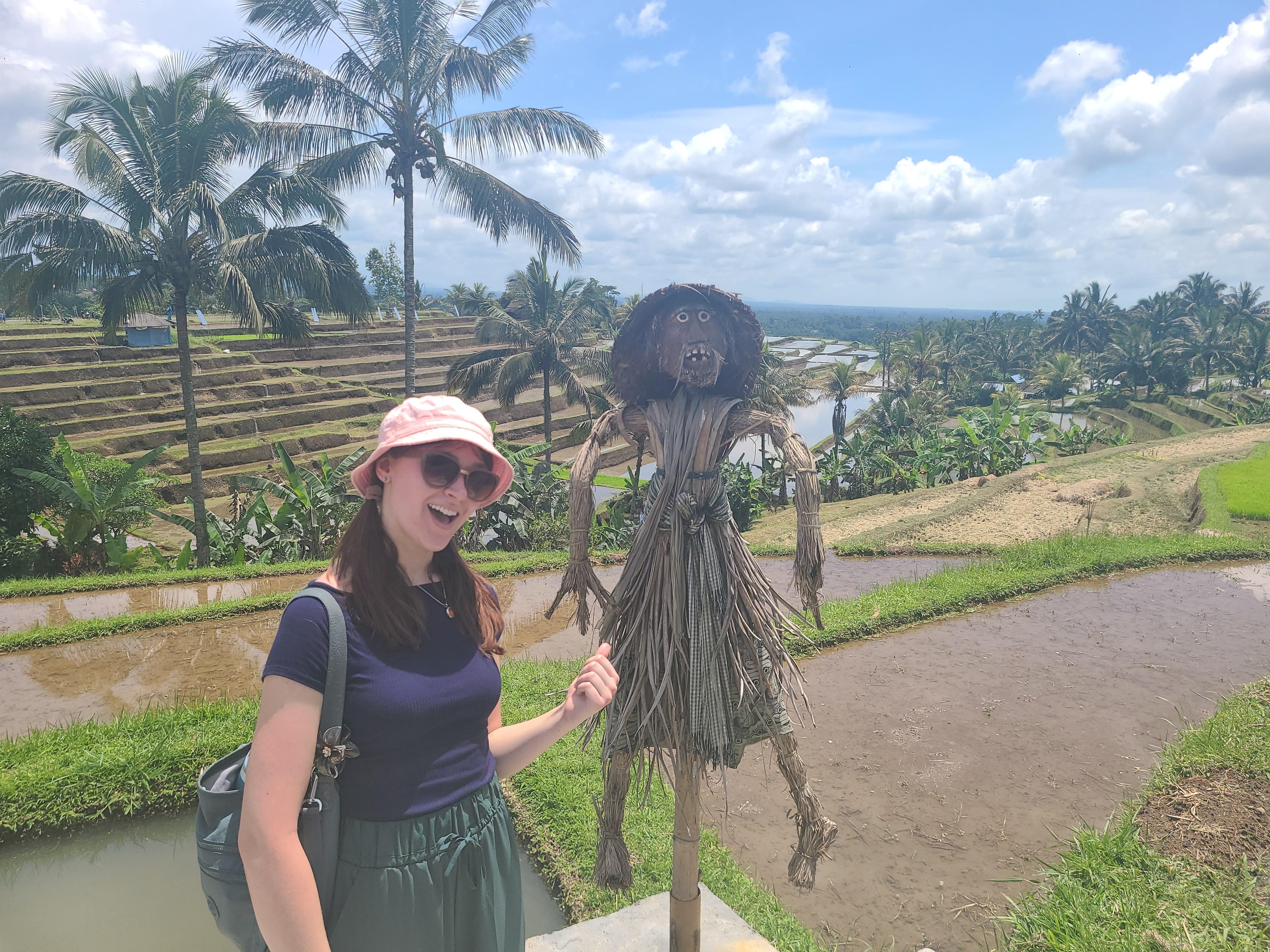 A student stands beside a traditional straw scarecrow overlooking terraced rice fields and palm trees under a bright blue sky.