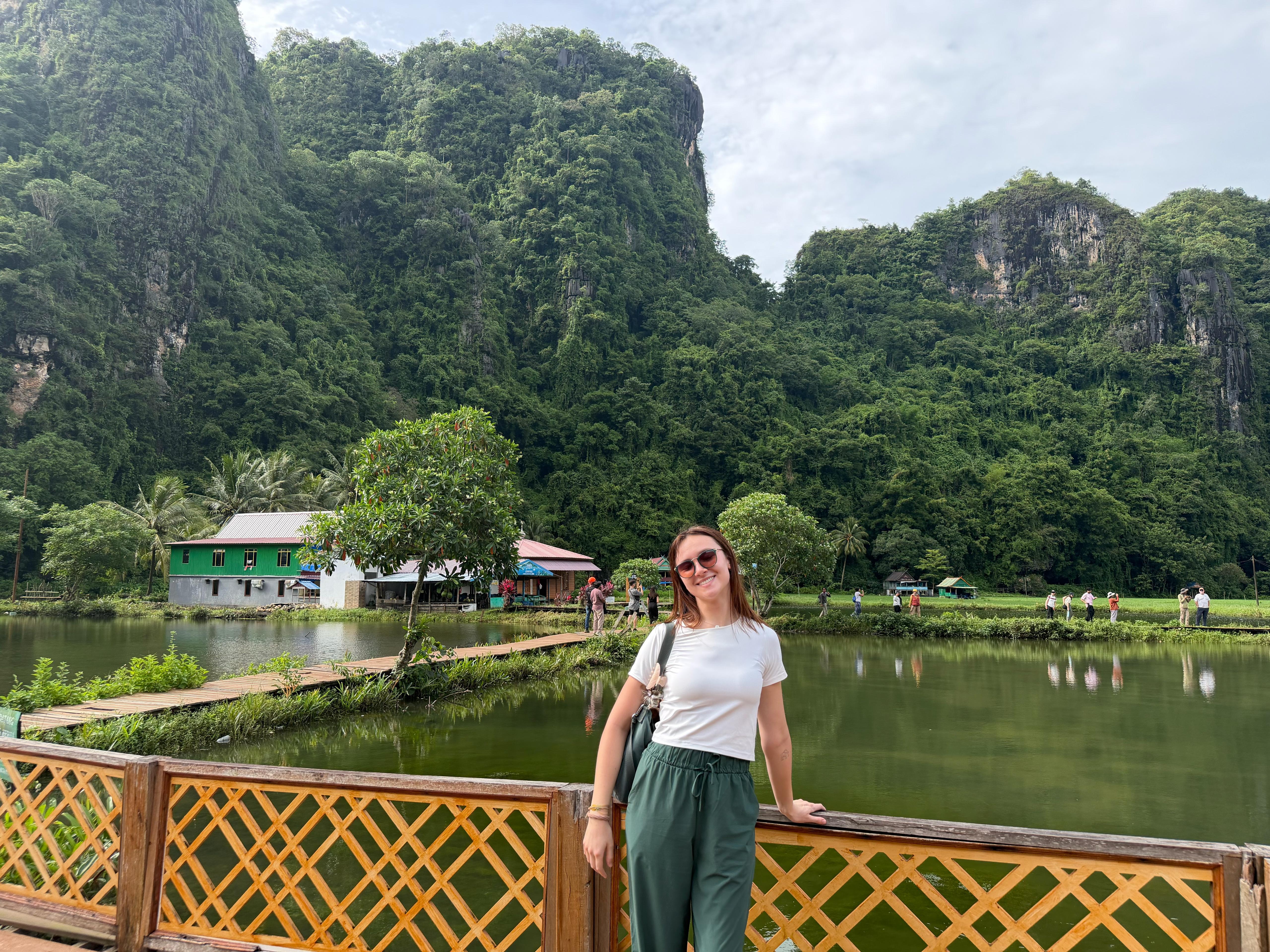 A student poses by a green lake with limestone mountains and small buildings in the background.