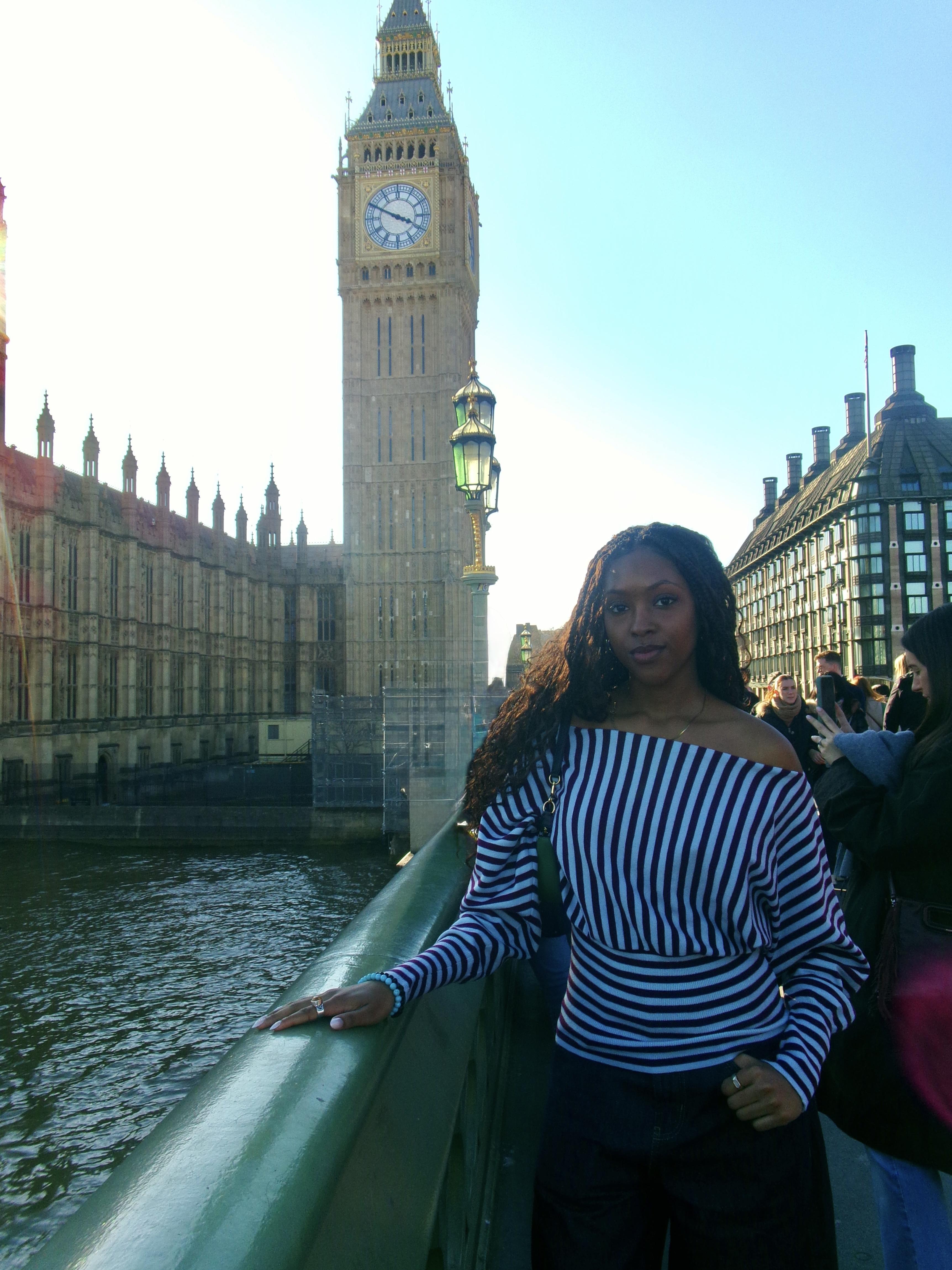 A student stands on a bridge over the River Thames in London with Big Ben and the Houses of Parliament in the background. She wears a striped off-the-shoulder top and rests one hand on the railing, with pedestrians and historic buildings visible along the river behind her.